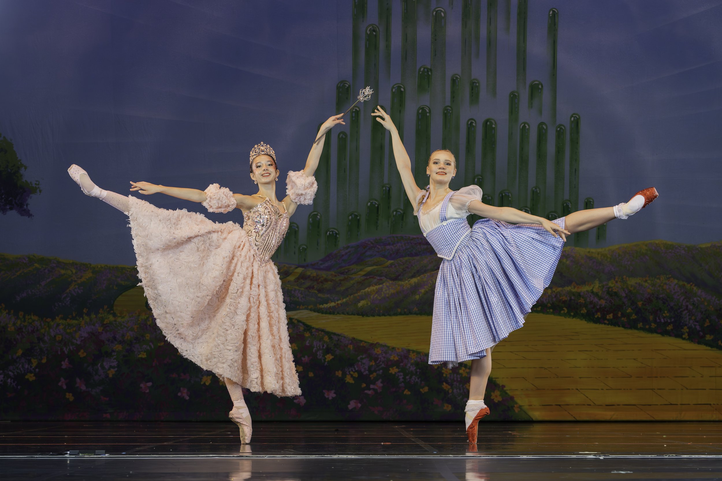 Two ballet dancers on stage performing a pose with a backdrop of a colorful landscape and green towers.