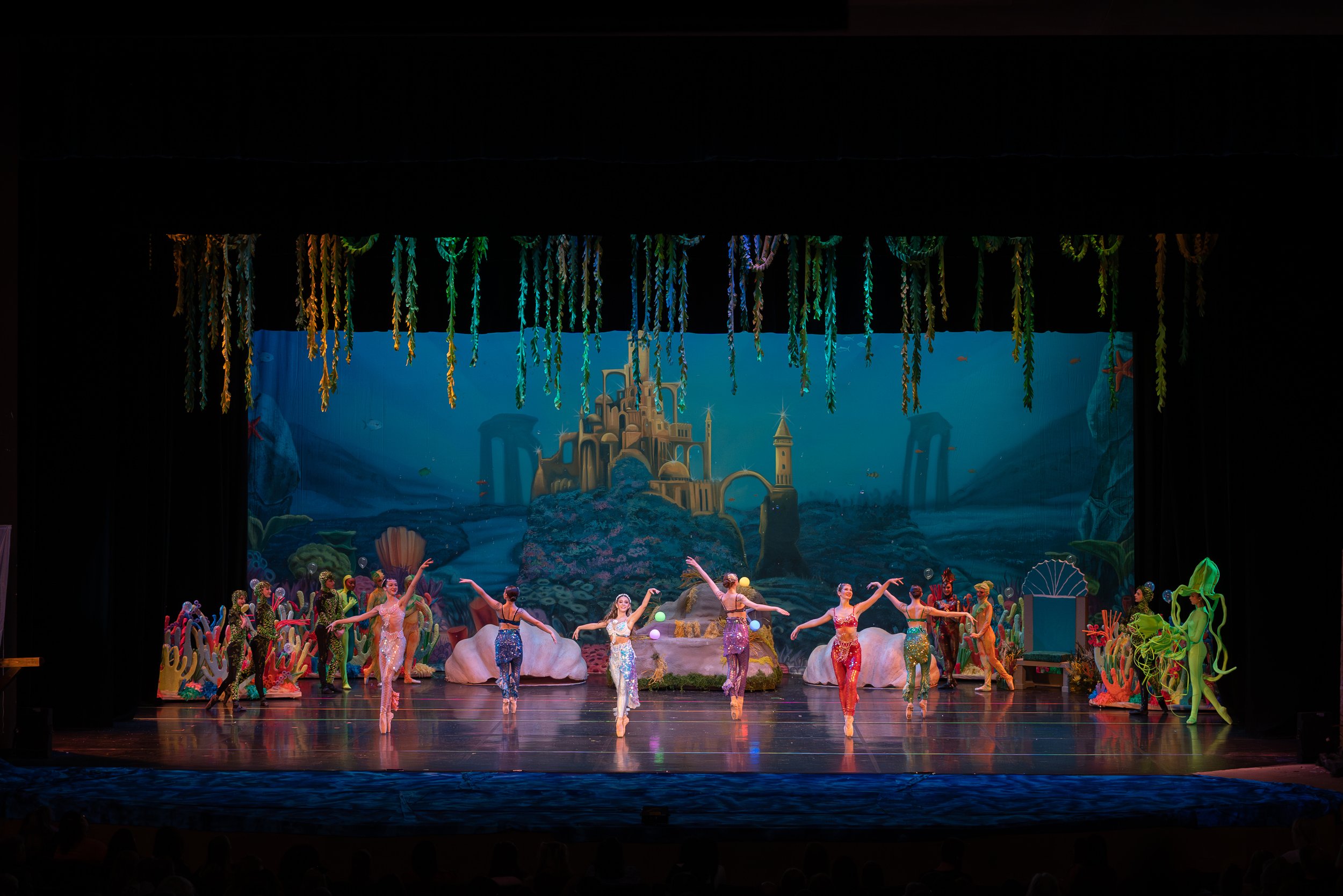Colorful stage performance of a ballet or dance show inspired by Under the Sea theme, with dancers in vibrant costumes and a scenic backdrop of an underwater castle and ocean scene.
