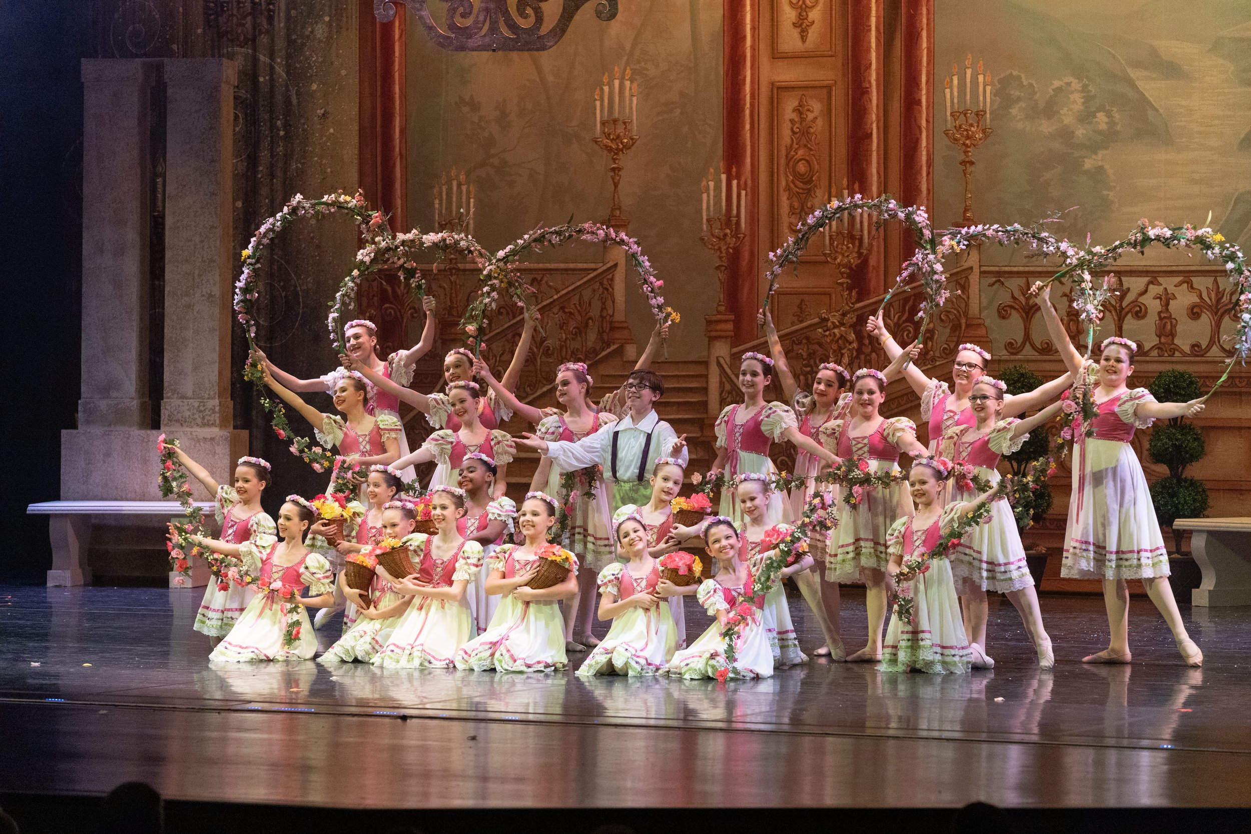Group of young ballet dancers in floral costumes performing on stage with a backdrop of a painted scene and wooden architecture, some holding flower baskets and floral hoops.