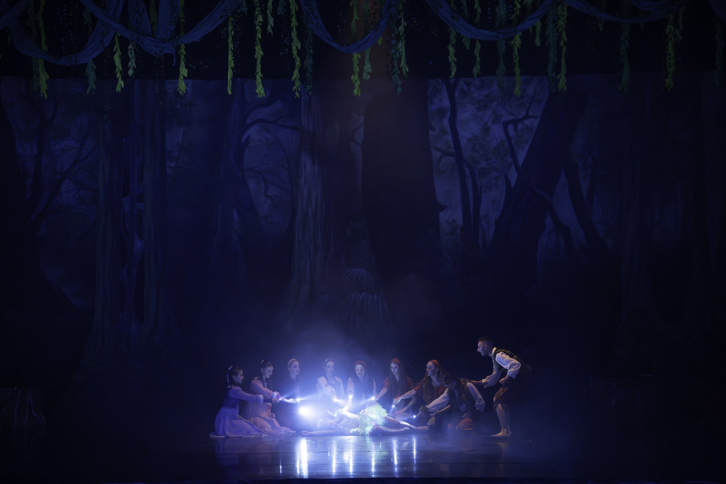 A theatrical stage scene depicting a group of performers surrounding a glowing object, with a backdrop resembling a dark, mystical forest with hanging vines and large tree silhouettes.