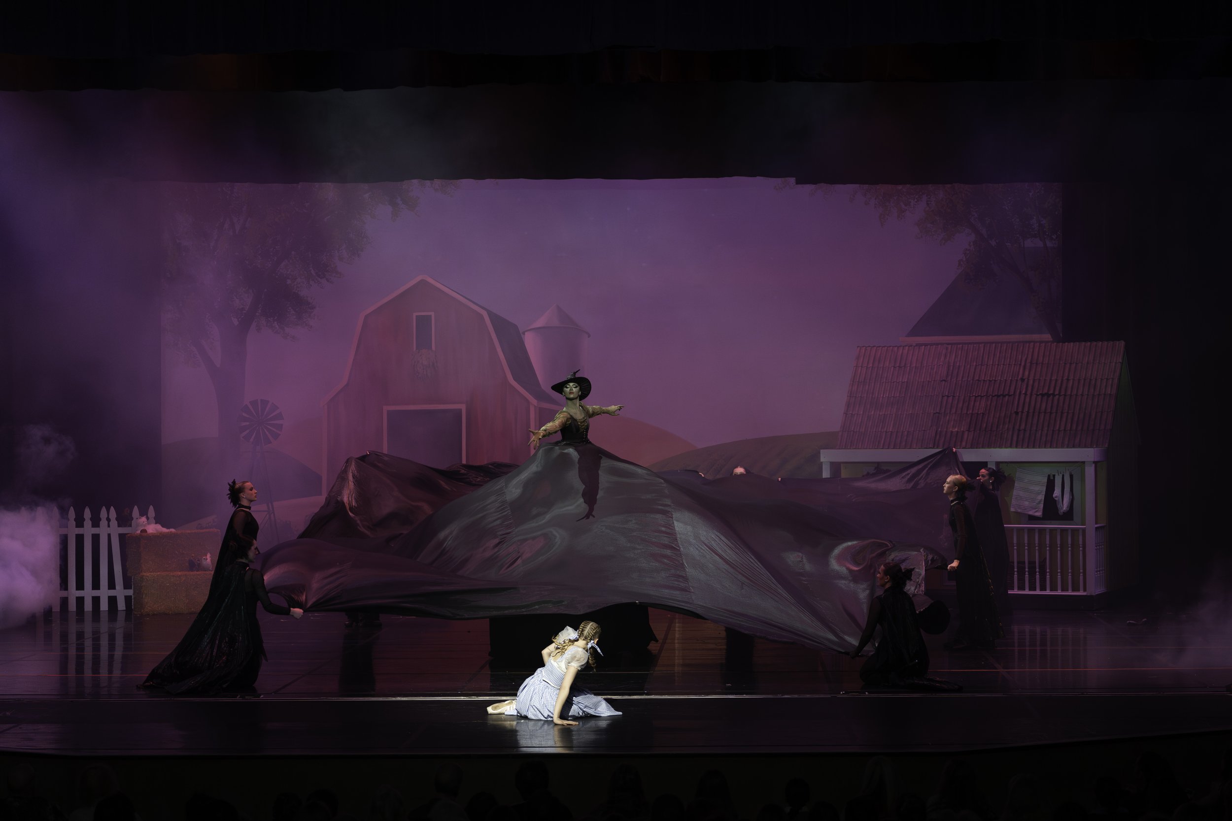 Stage scene from a theatrical performance with actors and a backdrop of a farm scene, featuring a woman in a large skirt acting as a hill and others in black costumes.