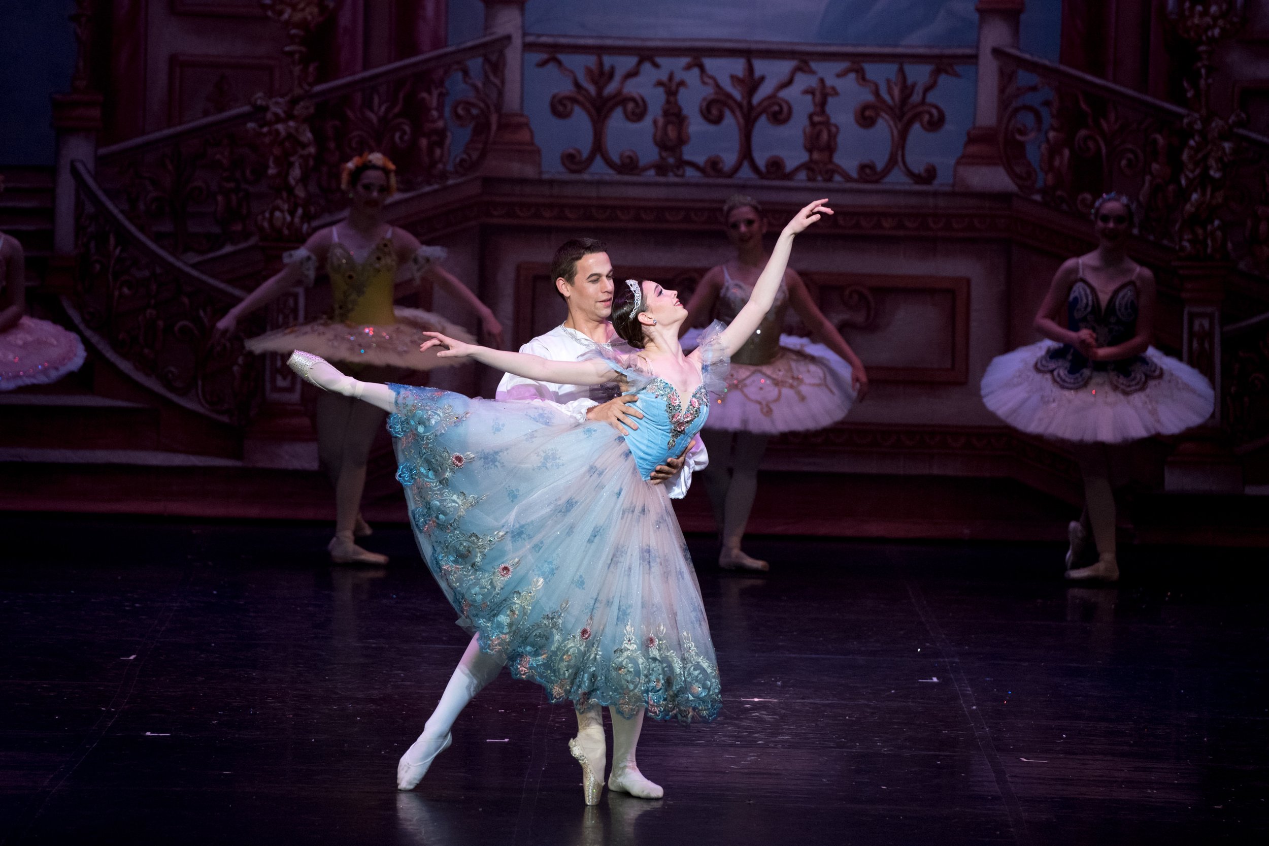 Ballet dancers performing on stage, with a female dancer in a blue tutu and a male dancer supporting her, surrounded by other ballerinas in tutus, in a grand theater setting.