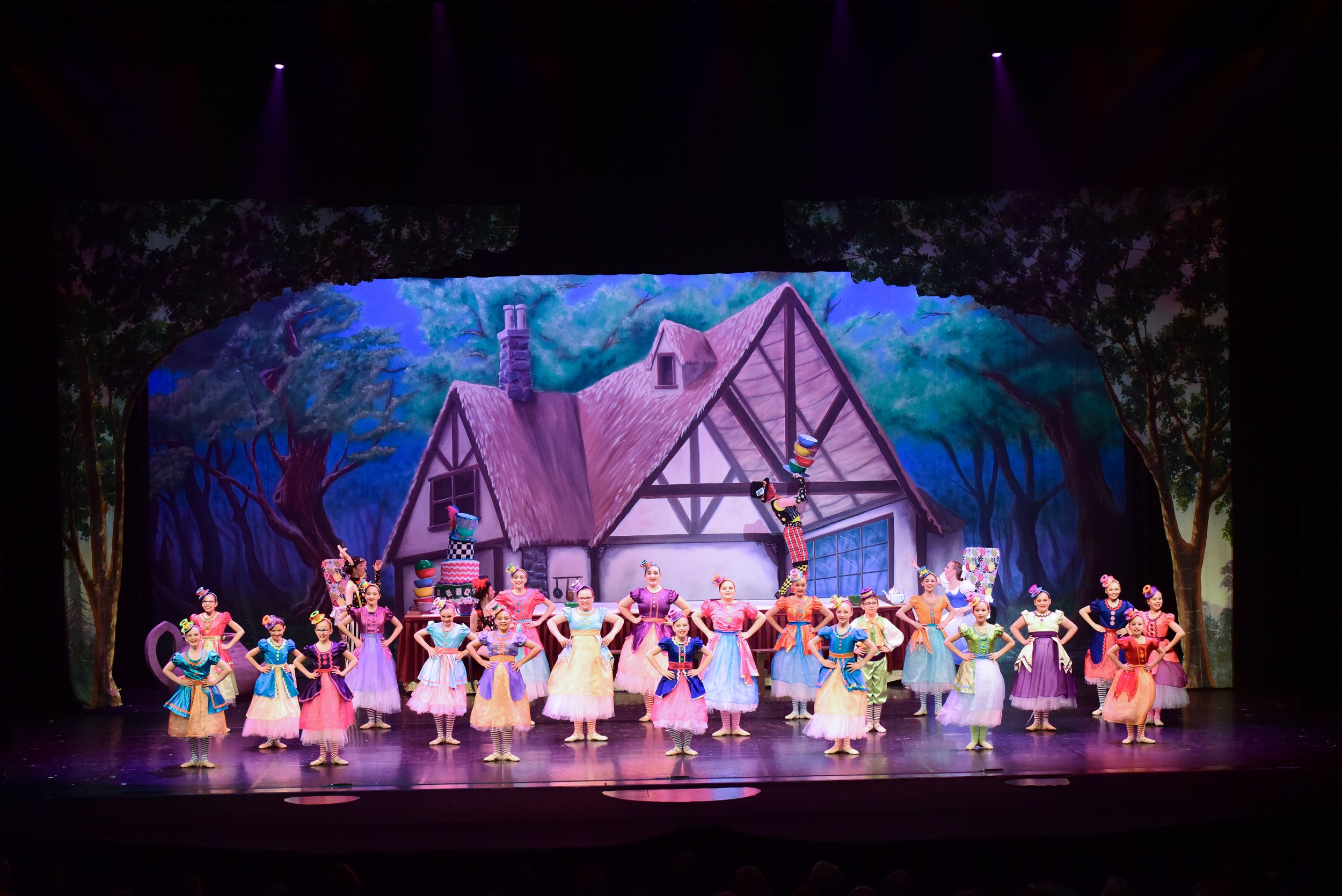 Theater stage with a colorful cast of dancers dressed in rainbow costumes performing in front of a painted woodland and house backdrop.
