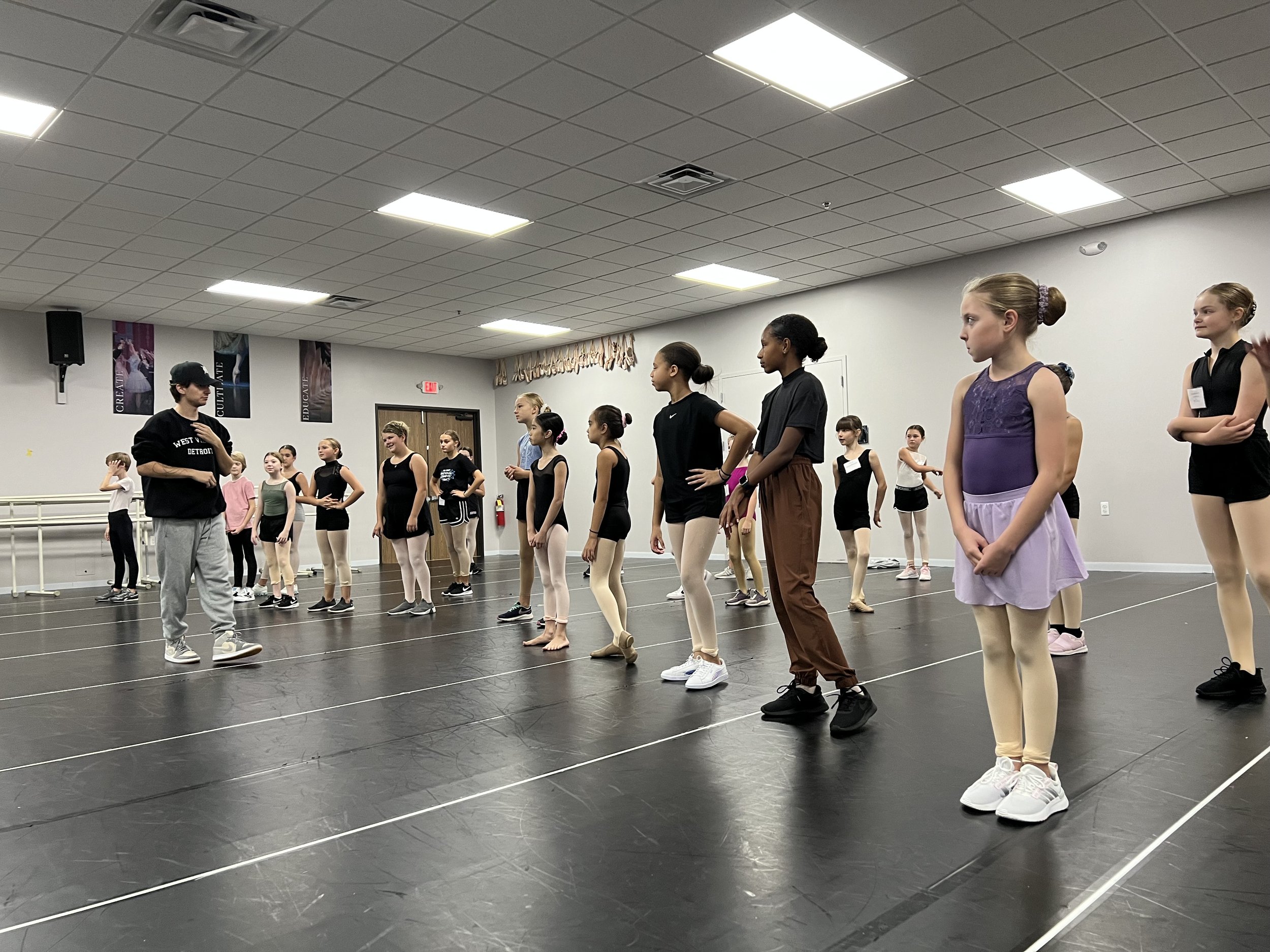 A group of young ballet students standing in a dance studio listening to their instructor, with some wearing leotards and tights, and others in casual dance wear.