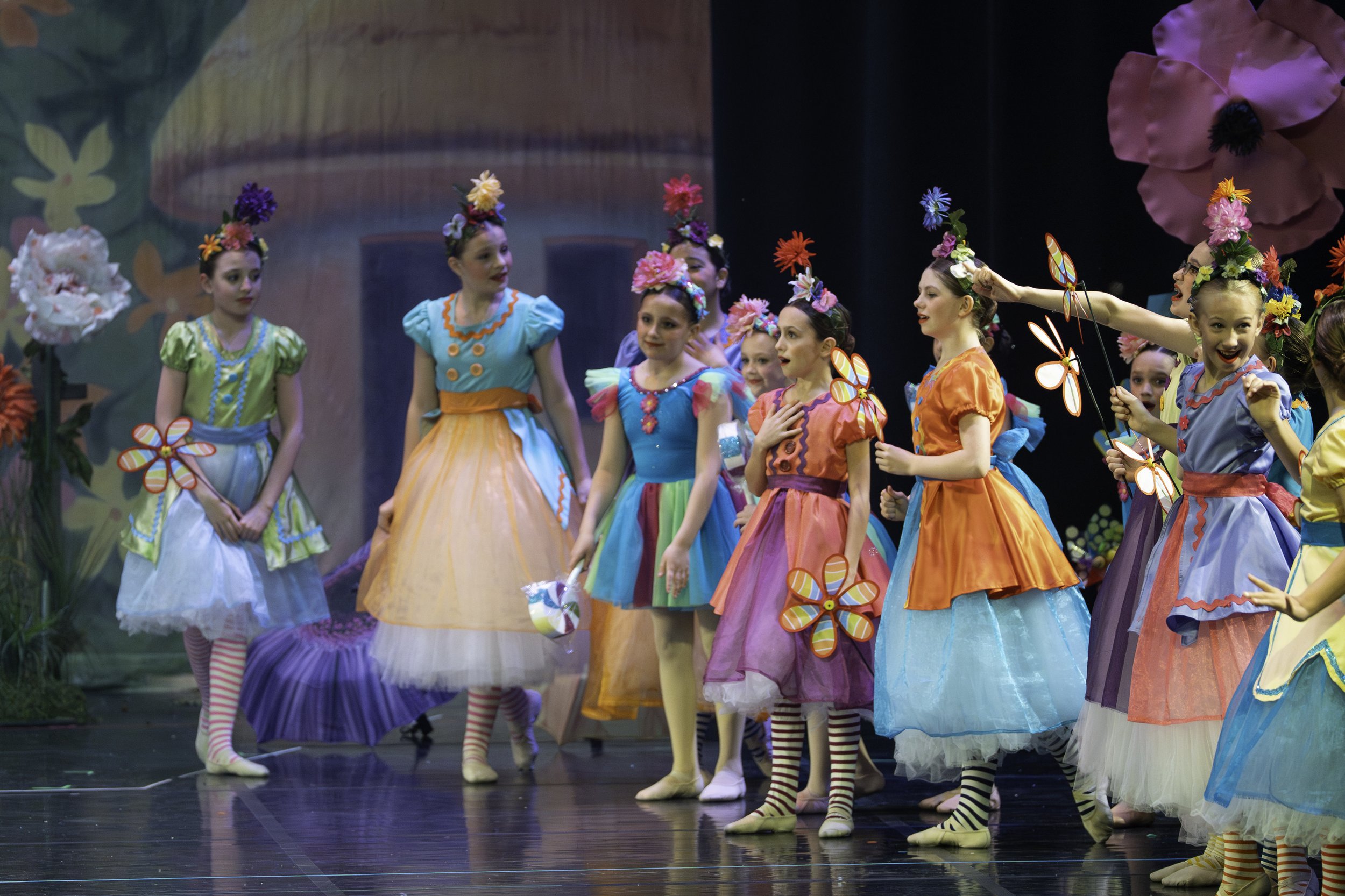 Young girls dressed in colorful costumes and flower headpieces performing on stage with a painted backdrop.