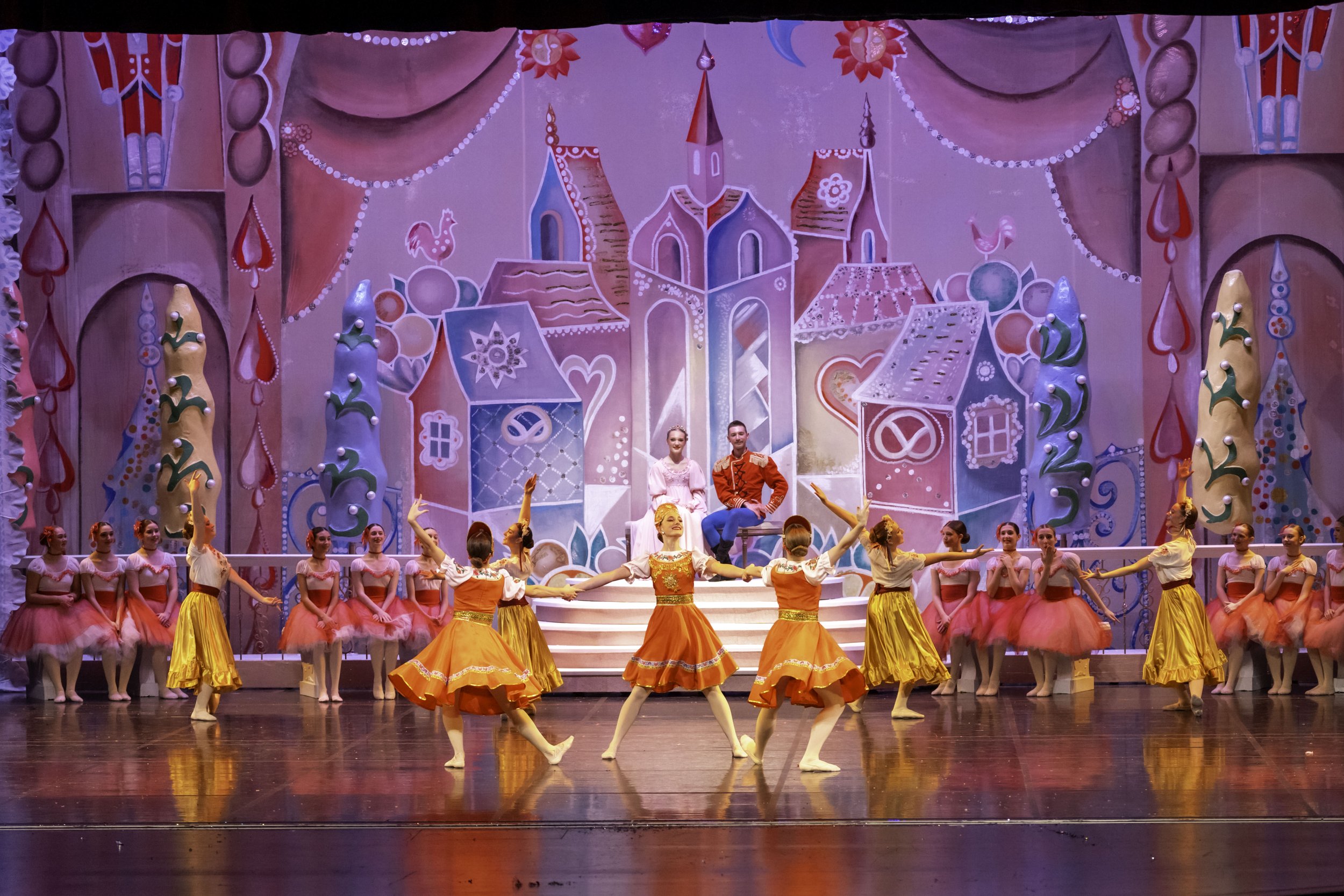 A stage performance of a ballet with dancers in colorful costumes, set against a backdrop of a whimsical, fairy-tale castle and garden scene.