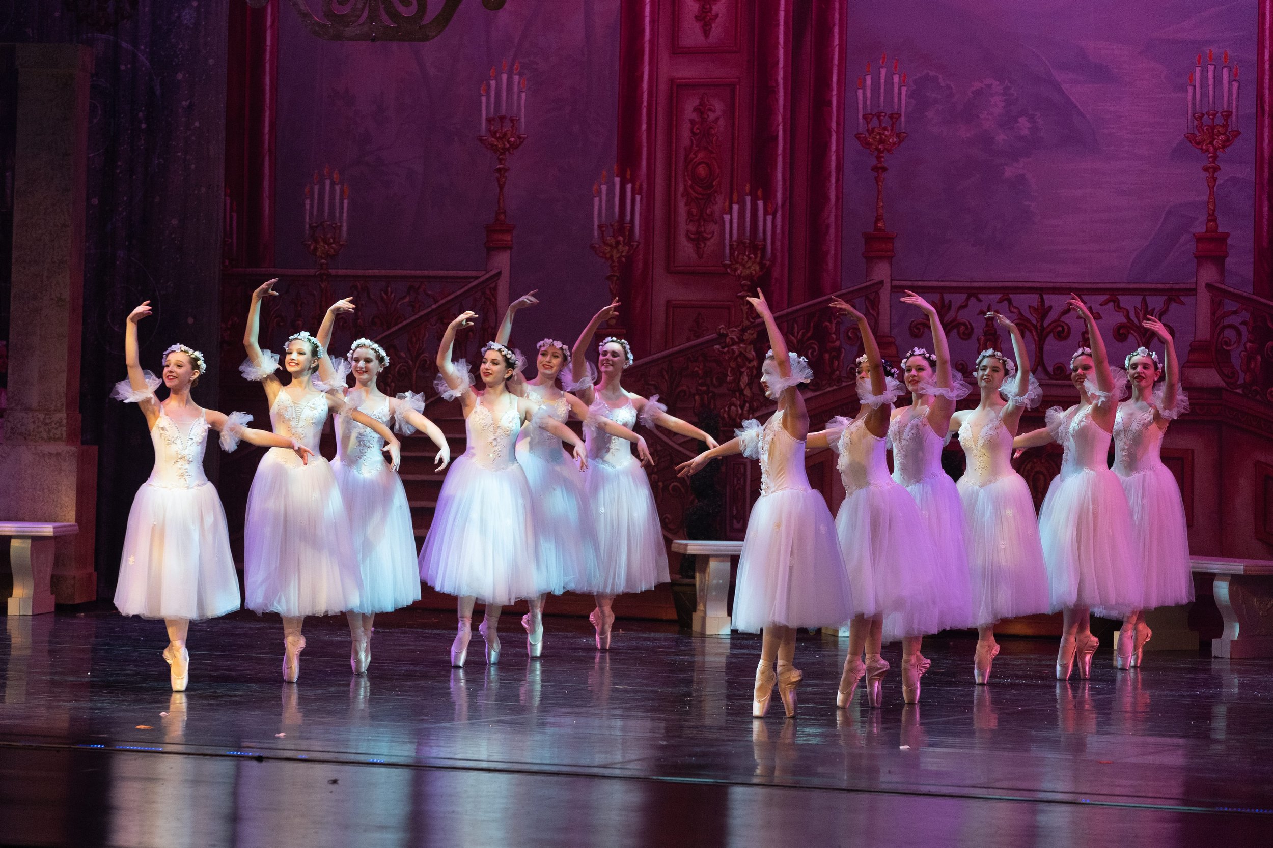 A group of ballerinas in white tutus performing on stage with pink lighting and ornate backdrop.