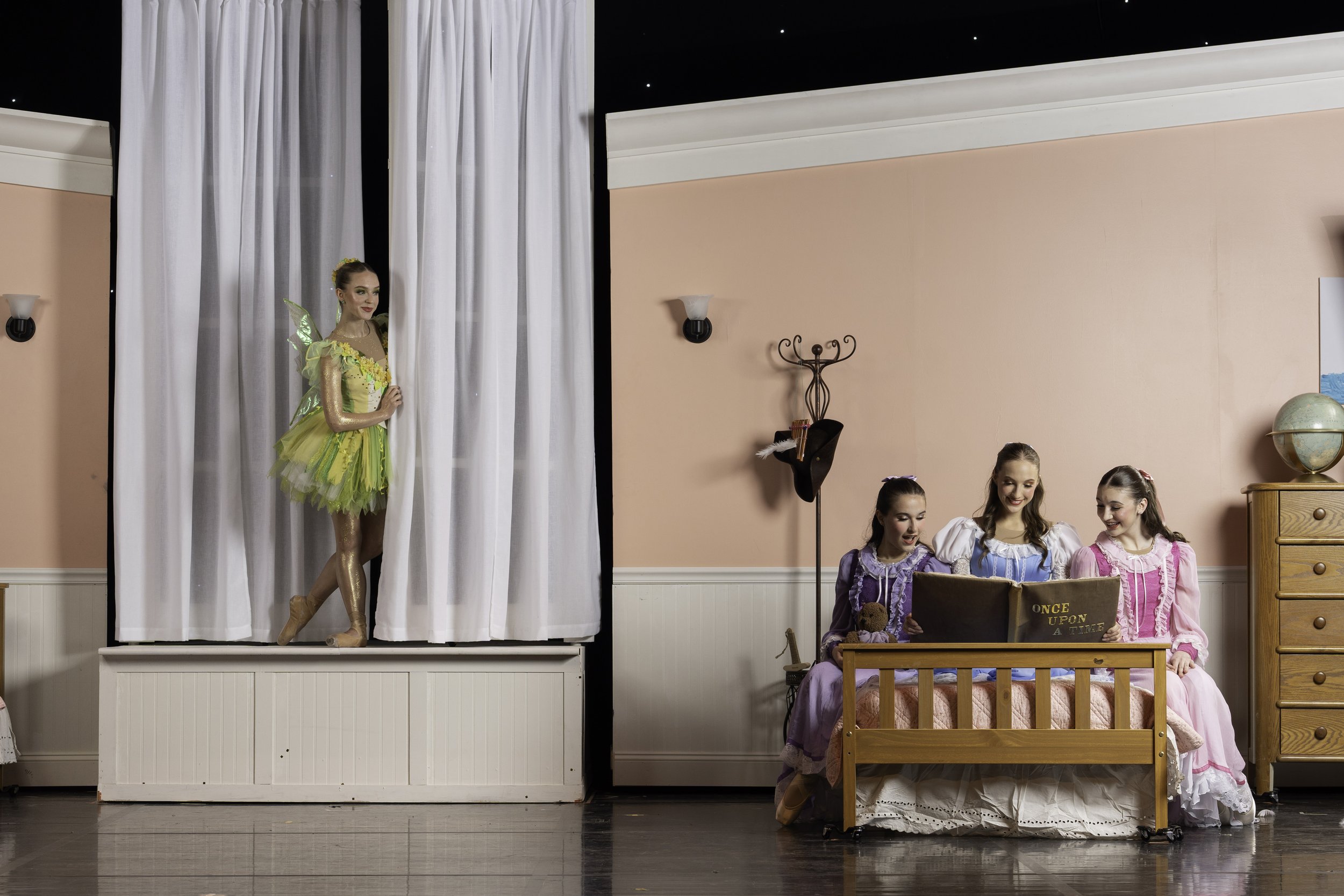 Three girls dressed as fairy tale characters sitting on a bed, reading a book titled 'Once Upon A Time,' while a girl dressed as a fairy or princess peeks out from behind curtains on a stage.