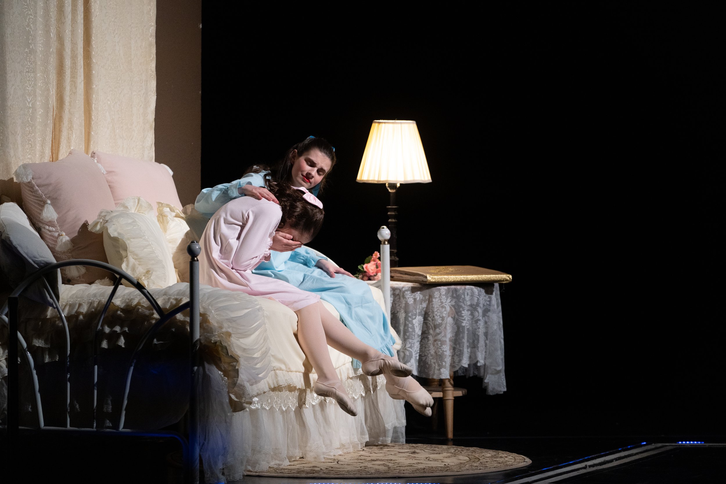 Two women sit on a bed with cream-colored ruffled bedding, one comforting the other who is crying, in a dimly lit room with a lamp on a side table.