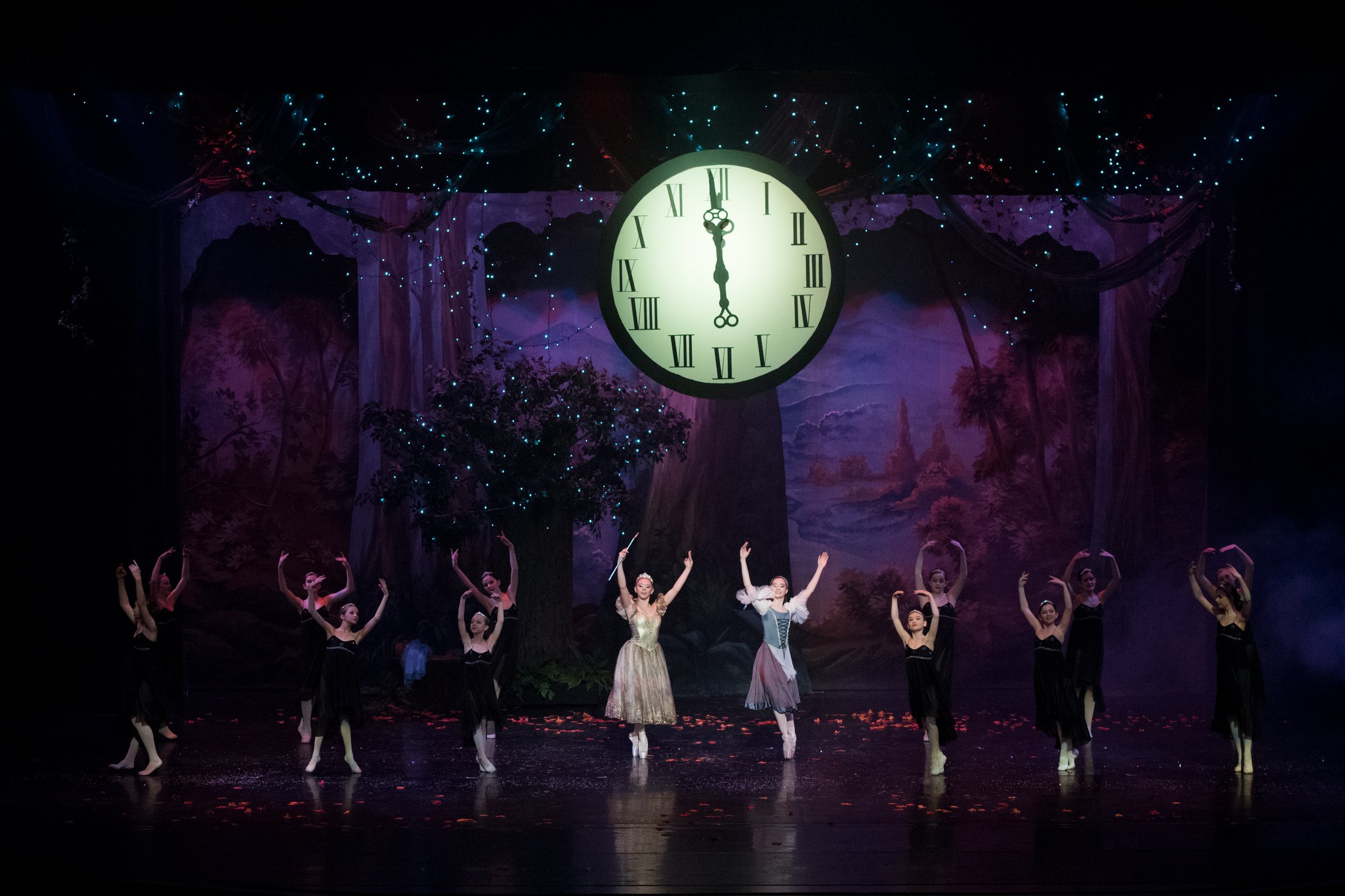 Theatrical stage with ballet dancers performing in front of a backdrop featuring a large clock showing midnight, trees, and a starry night sky.