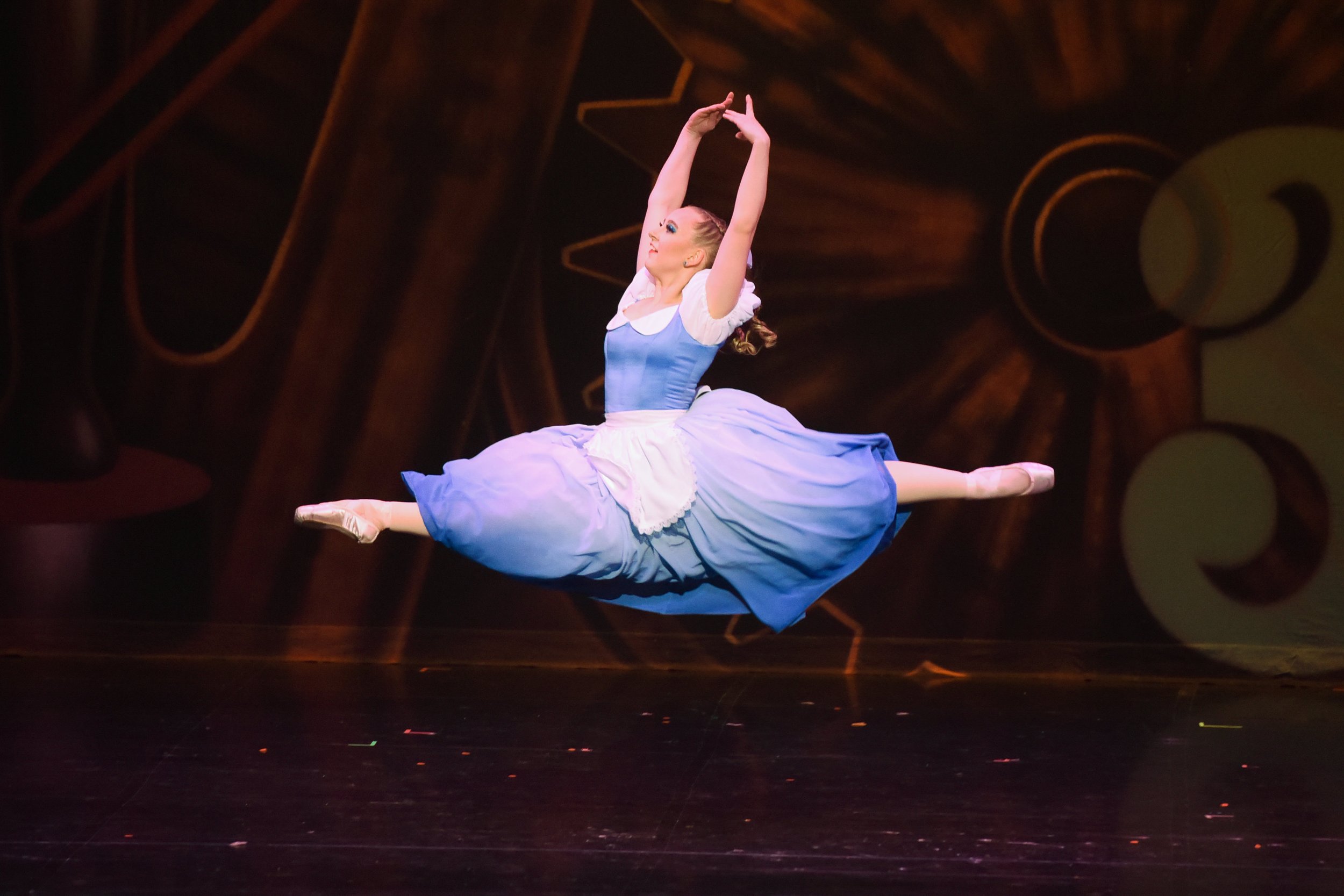 A ballet dancer in a blue dress performing a jump on stage with a dark, theatrical backdrop.