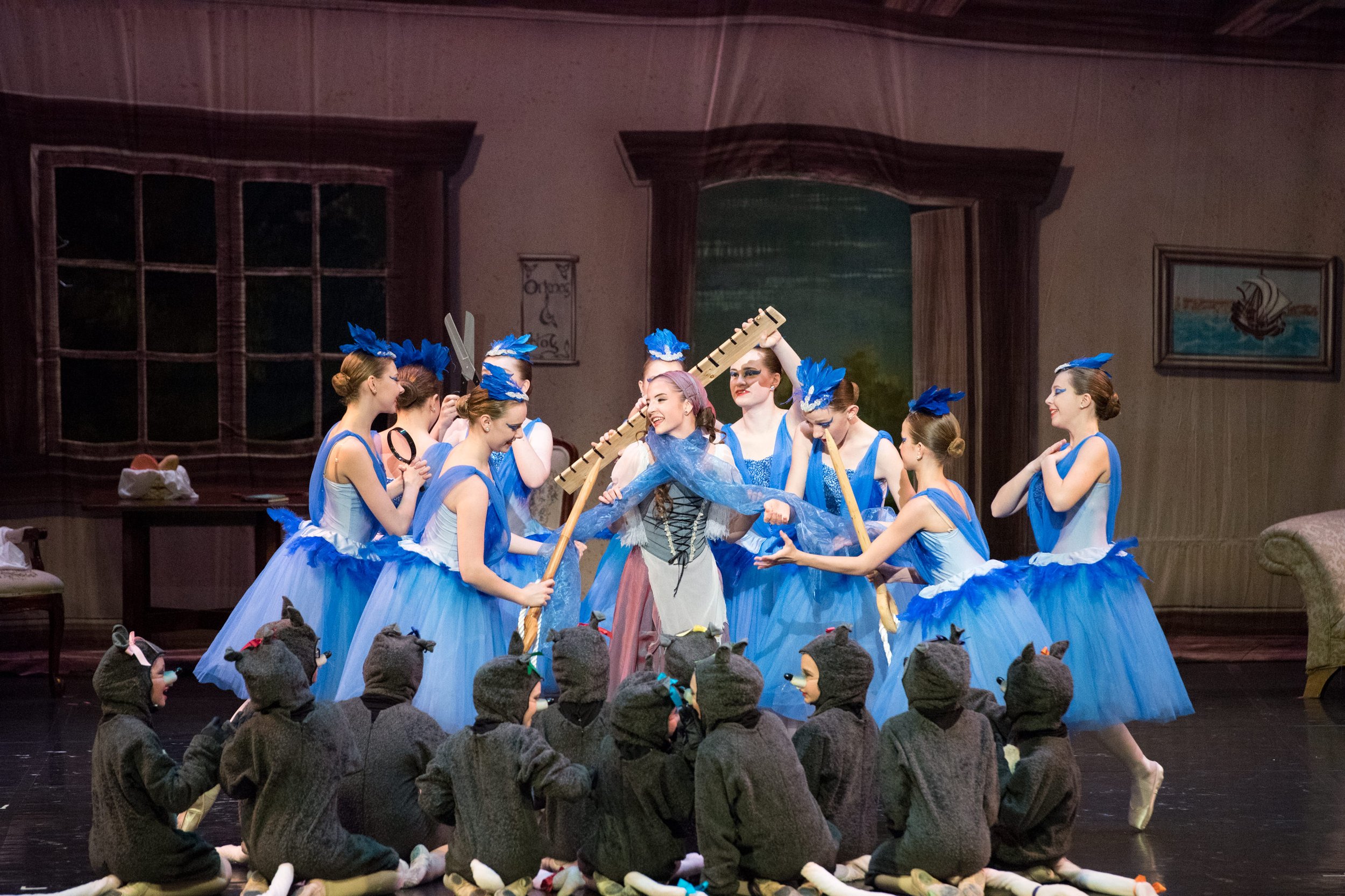 Ballet dancers in blue tutus performing on stage with children in animal costumes seated in front. The scene is set in a living room with painted windows, a door, and a painting of a ship.