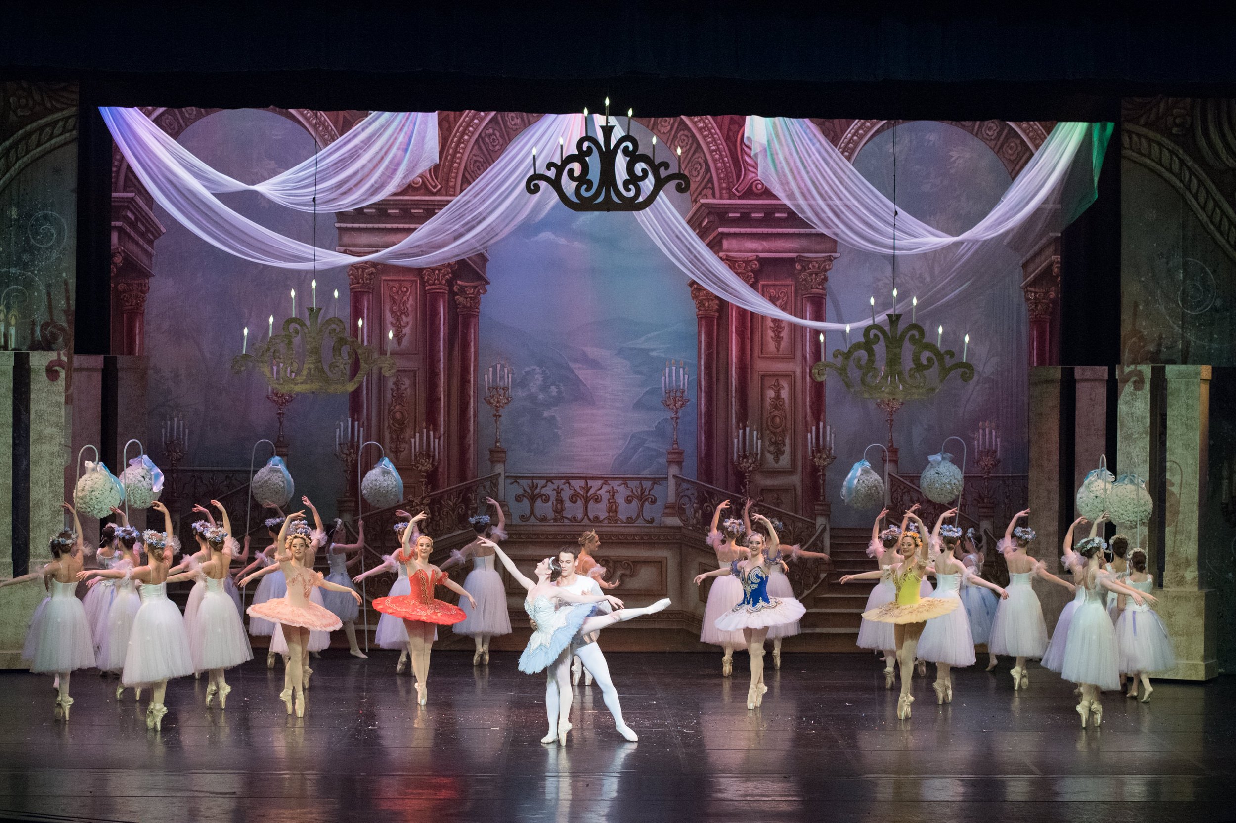 Ballet dancers performing on stage with a backdrop of a grand, ornate theater interior, featuring chandeliers, draped fabric, and a painted landscape. The dancers are in colorful tutus, with some holding flower baskets.