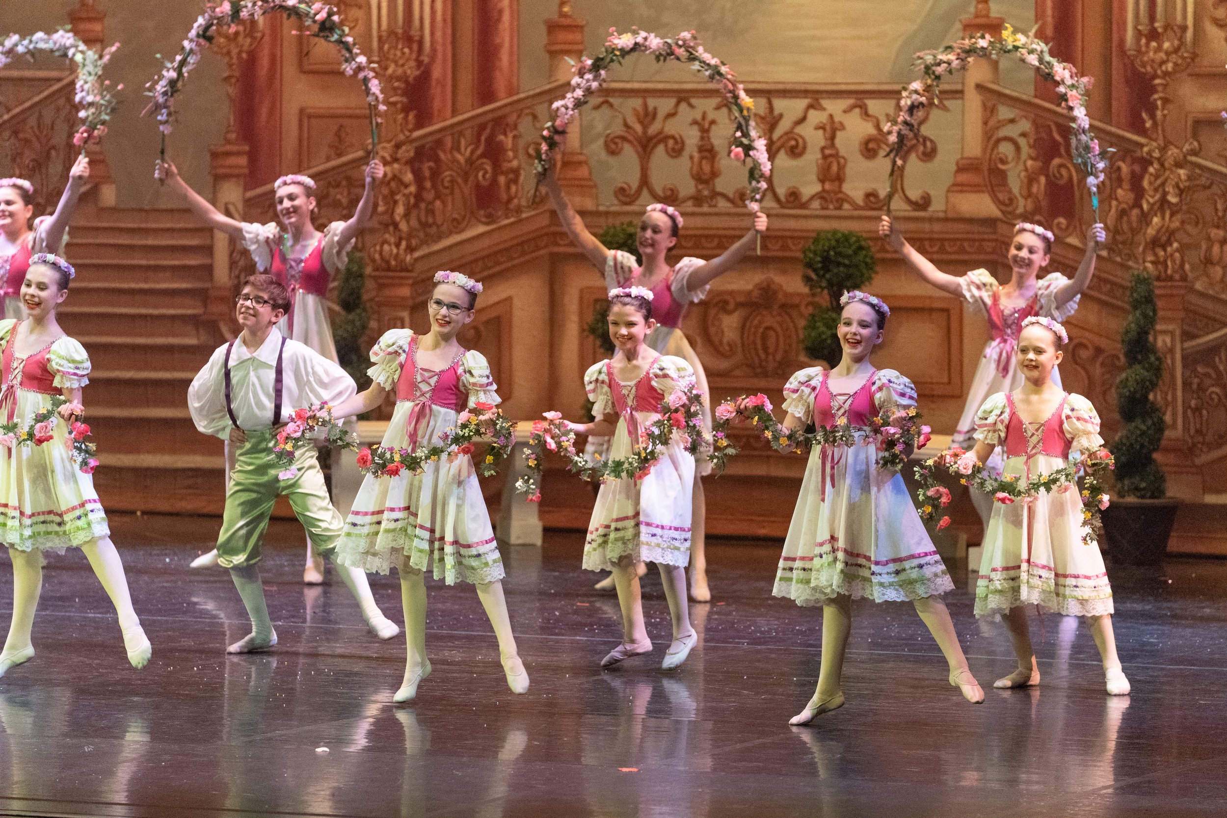 Children performing a ballet on stage, wearing pink and cream costumes with floral accents, with a decorated wooden staircase and floral arches in the background.