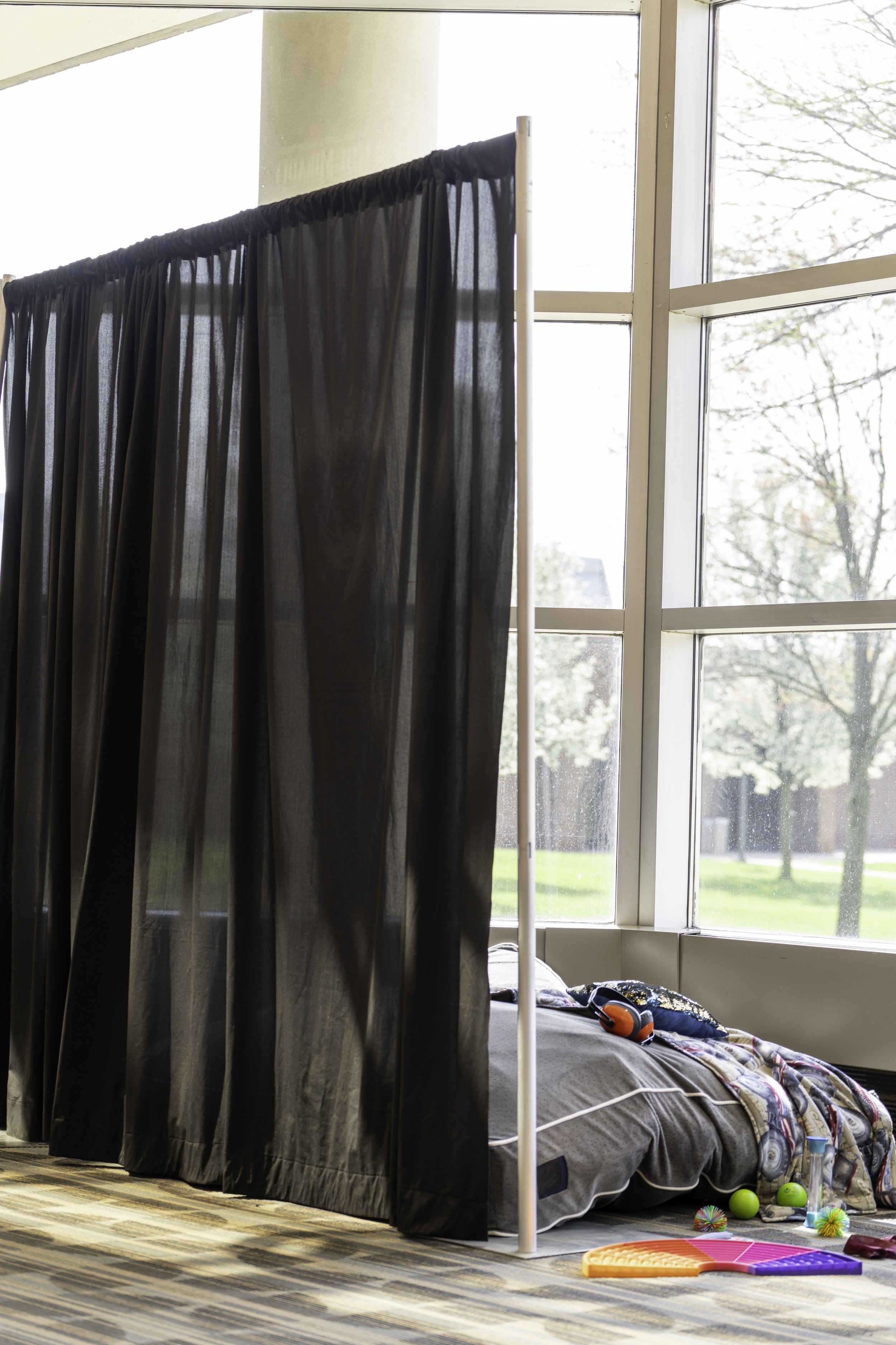 A makeshift bed on the floor next to a large window with black curtains, surrounded by toys such as balls, puzzles, and plush toys.