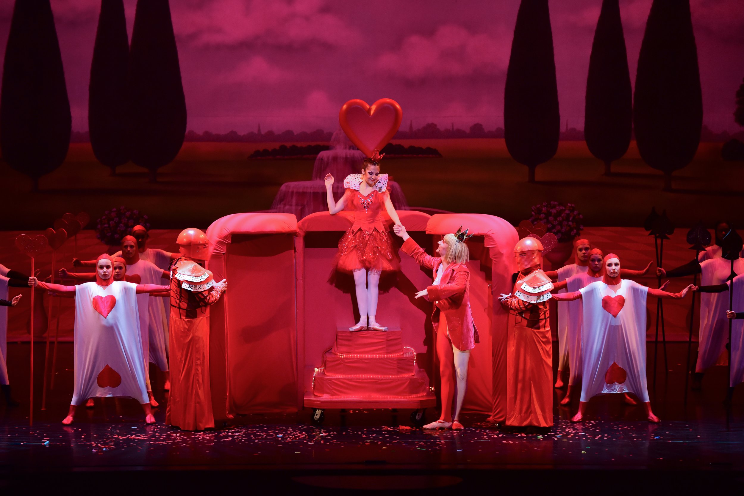 Theater production scene featuring characters dressed as playing cards, with a woman in a red dress on a staircase, surrounded by dancers and a pink and red heart theme.