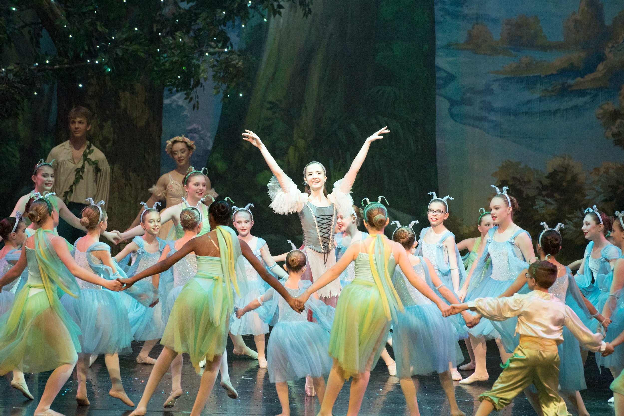 Ballet dancers dressed as insects performing on stage with a forest backdrop, including a woman in the center with arms raised, surrounded by young dancers holding hands in a circle.