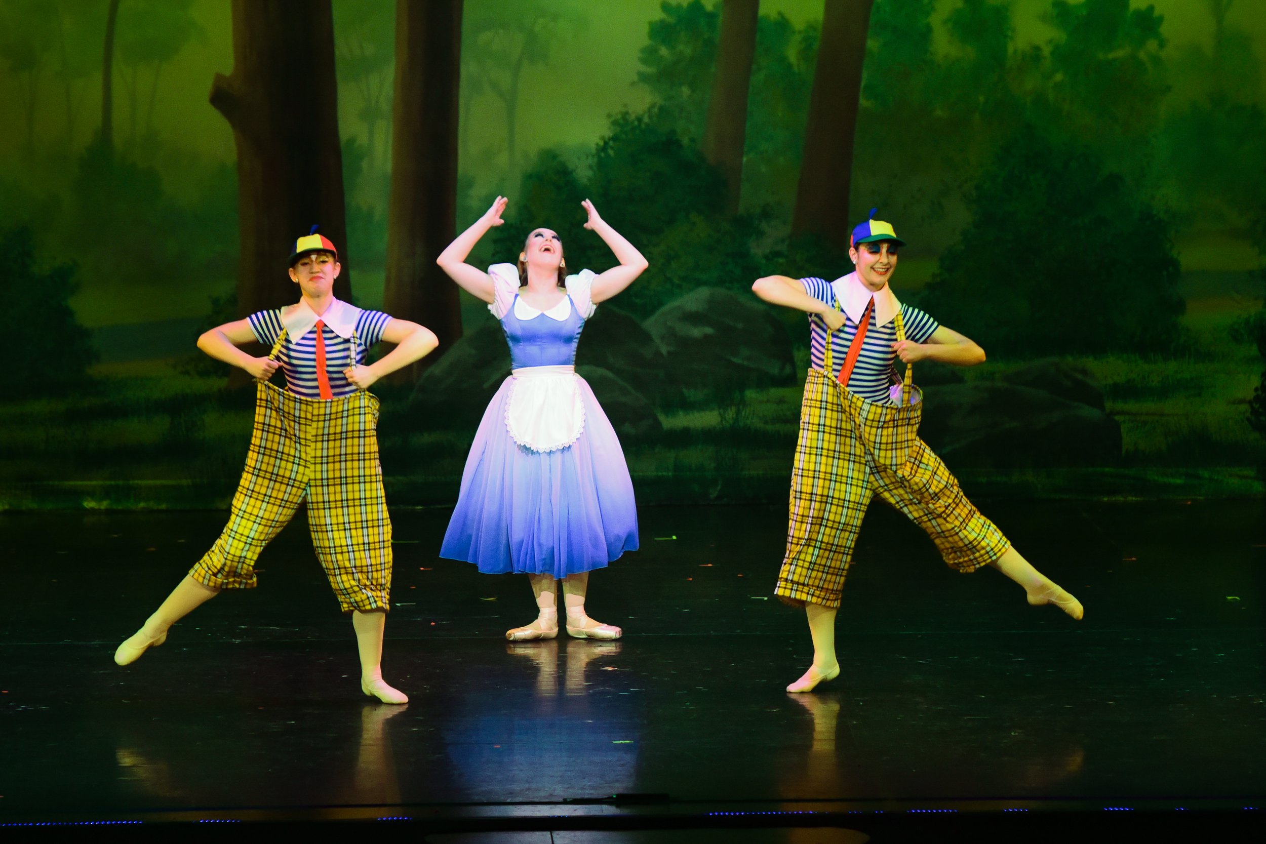 Three performers on stage in colorful costumes, with a forest backdrop. Two are dressed as puppeteers with yellow plaid pants and striped shirts, and one as a girl in a blue and white dress, likely portraying a scene from a theatrical production.
