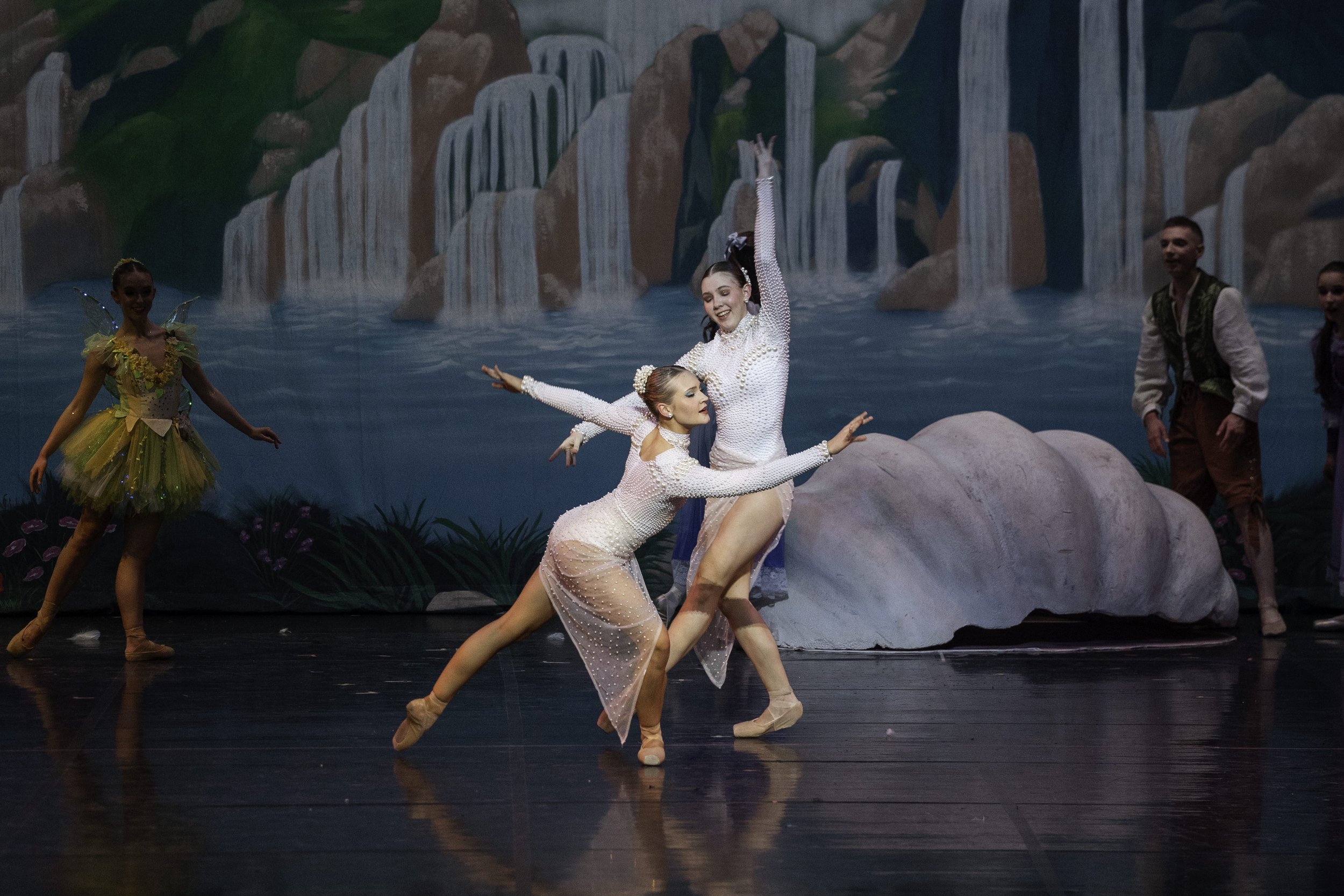 Ballet dancers performing on stage with a scenic backdrop of waterfalls and rocks, wearing costumes representing a fairy tale setting.