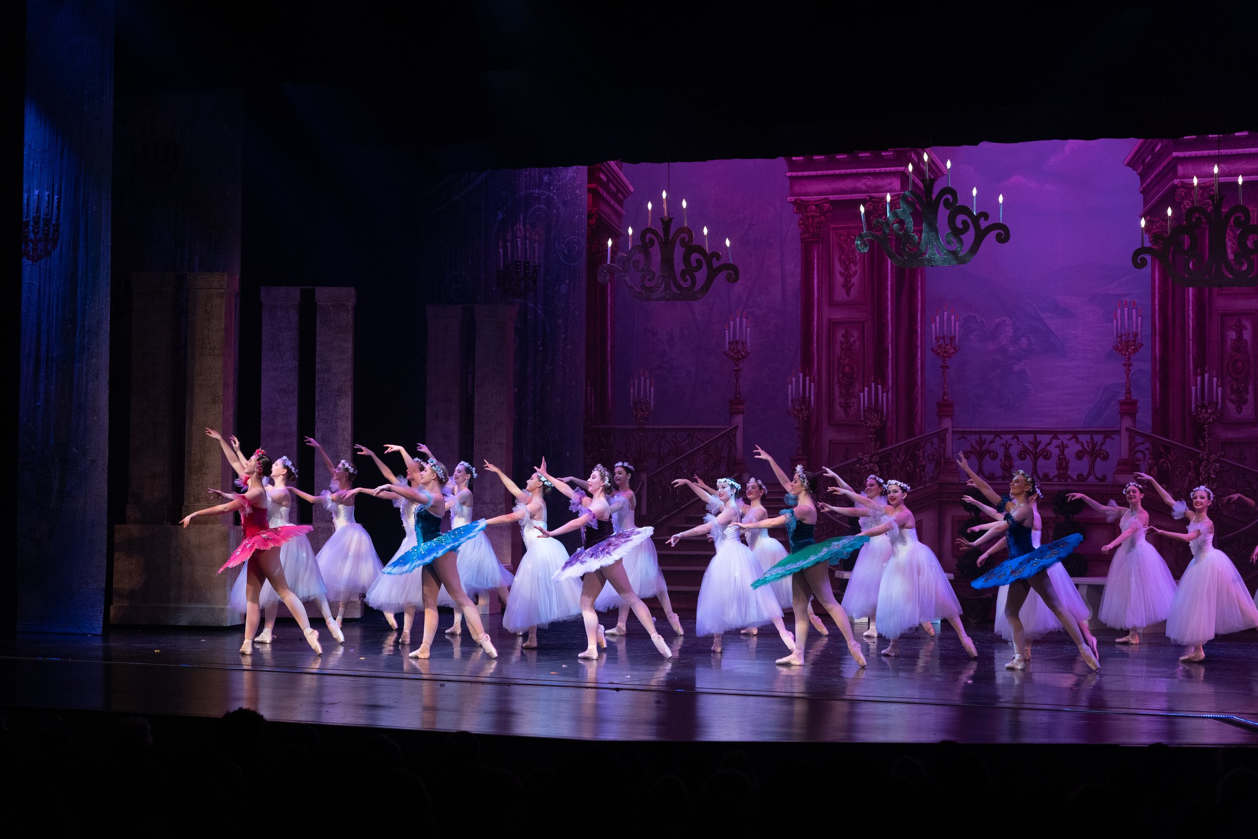 Ballet dancers performing on stage in colorful costumes with a theatrical backdrop and chandeliers.