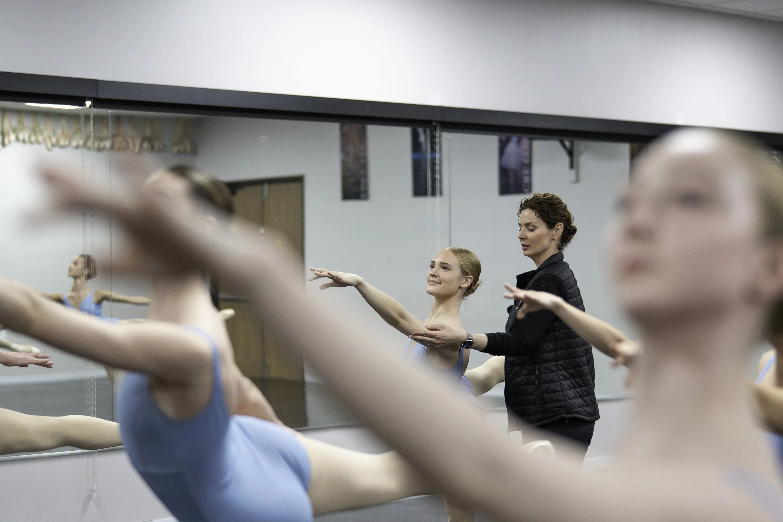 Ballet instructor helping young dancers stretch in a dance studio with a large mirror.