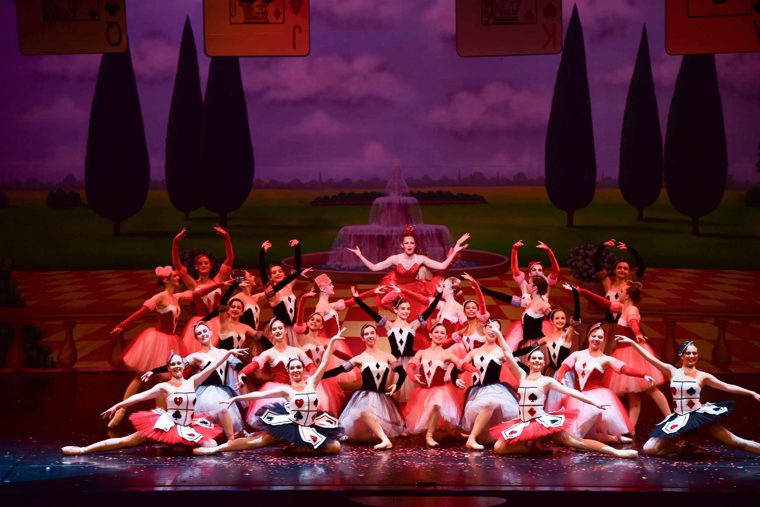 Ballet dancers performing on stage dressed as playing cards, with a backdrop of scenic countryside and a fountain.