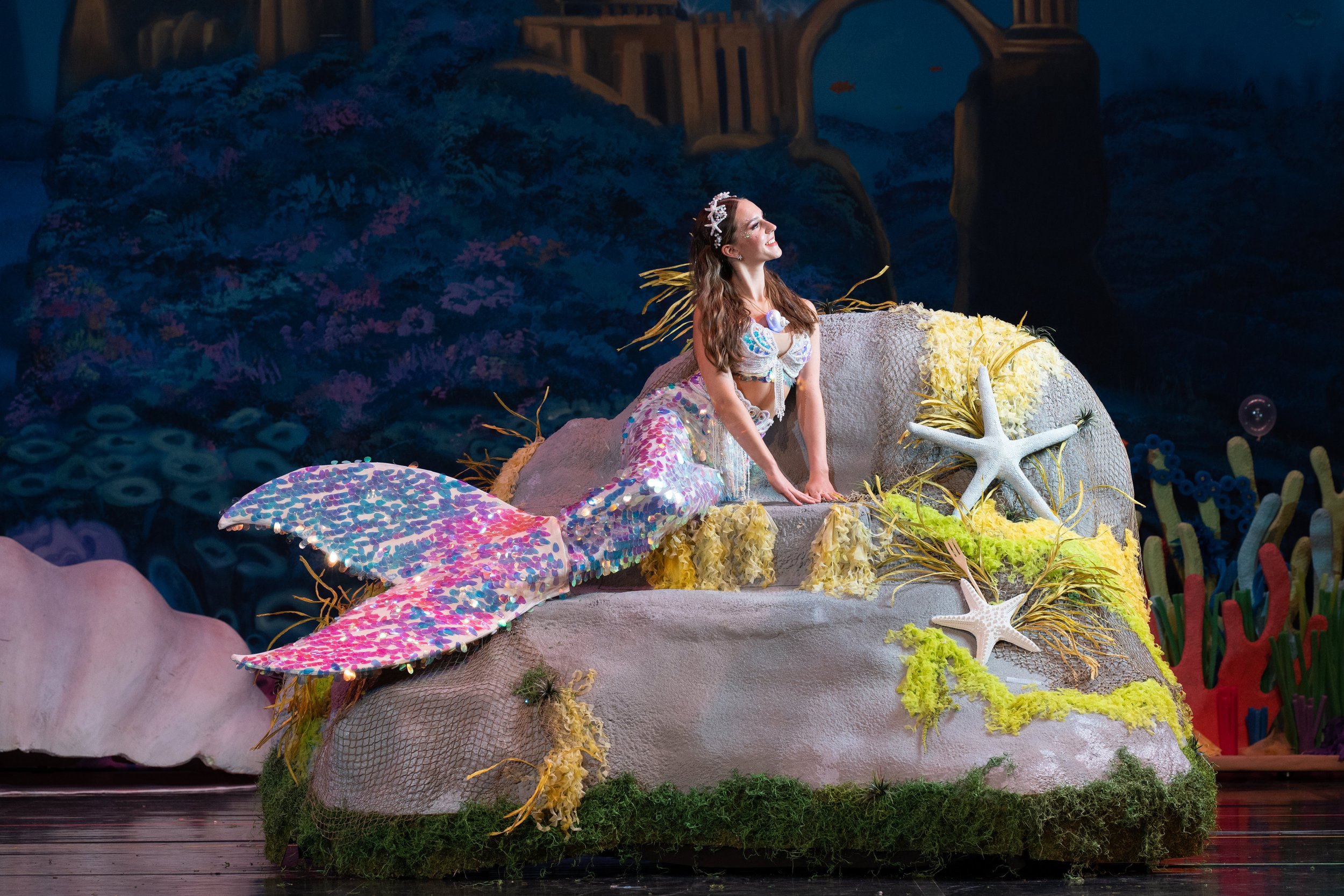 A woman dressed as a mermaid on a stage set with underwater-themed decorations, including starfish, seaweed, and coral, with a painted backdrop of the ocean.