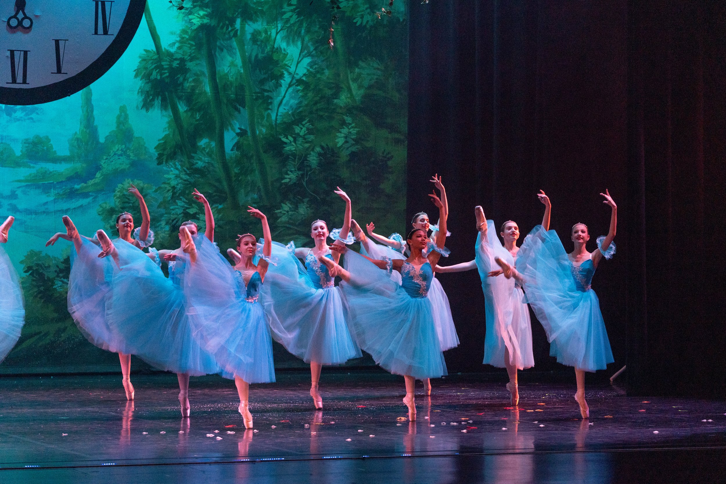 Ballet dancers in blue tutus perform on stage with a scenic backdrop of trees and a clock
