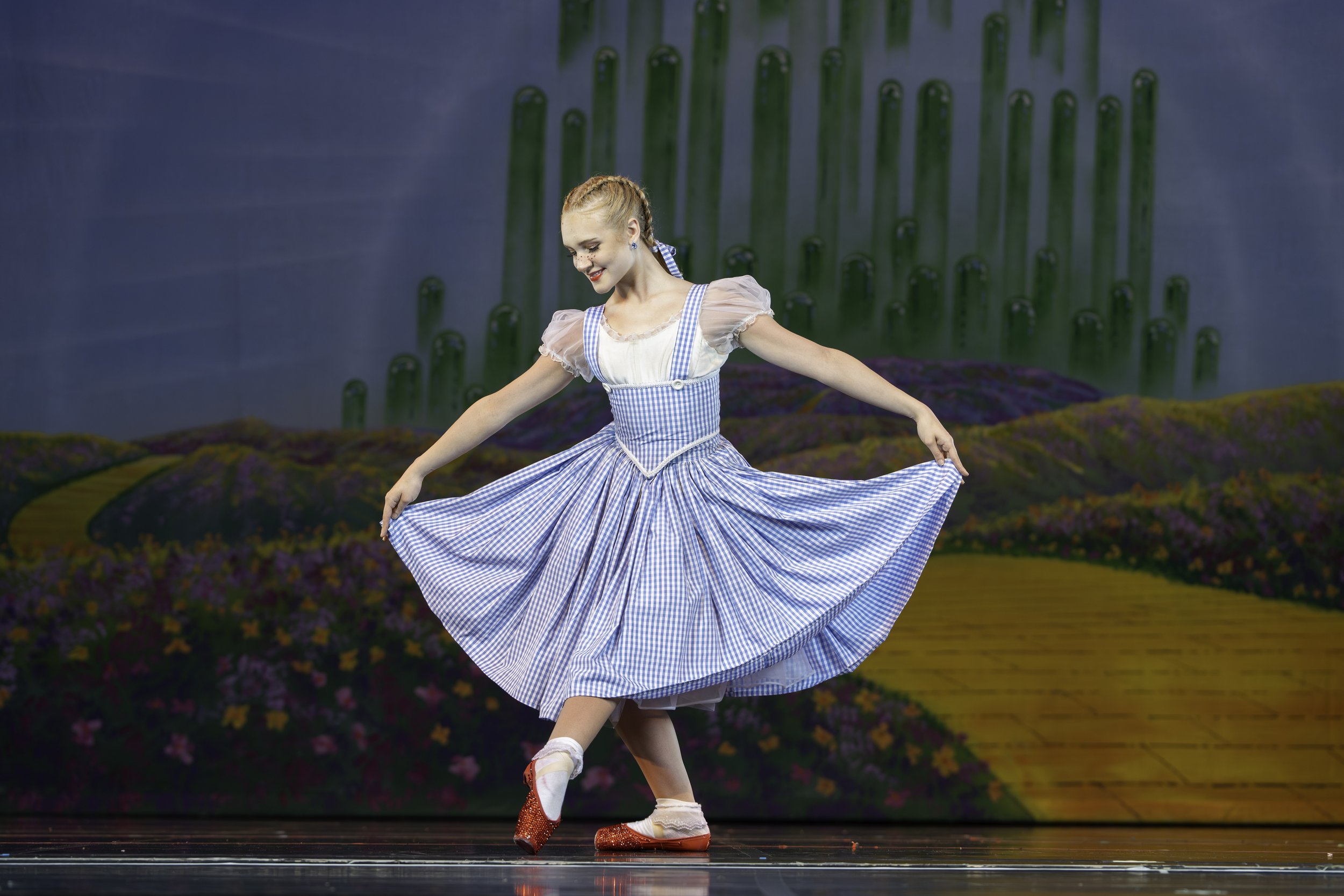 A young woman dressed as Dorothy from The Wizard of Oz, wearing a blue gingham dress and red shoes, performing a dance on stage with a painted backdrop of a yellow brick road, purple flowers, and green cacti.