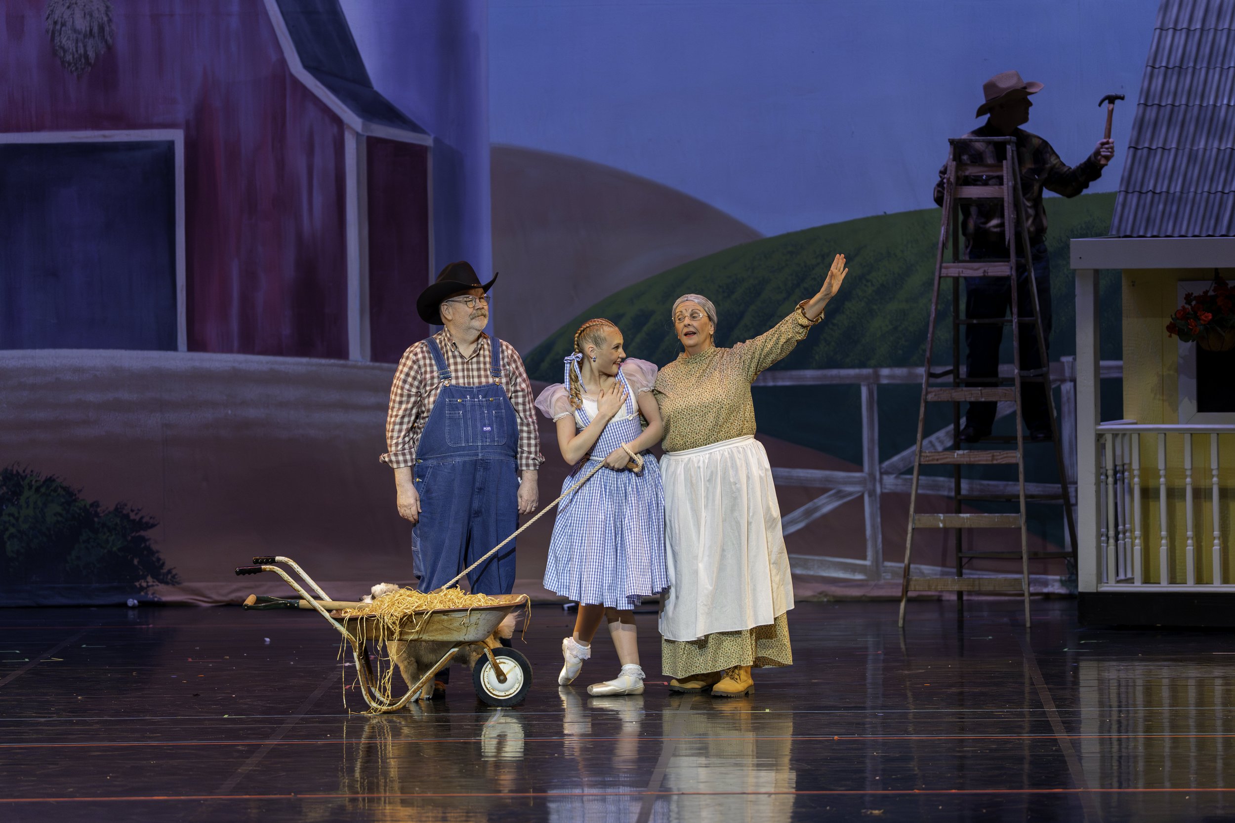 Actors performing a scene from a play on stage with farm set, including a man in suspenders and cowboy hat, a young girl in blue checkered dress, an older woman in a long skirt, and another actor on a ladder.