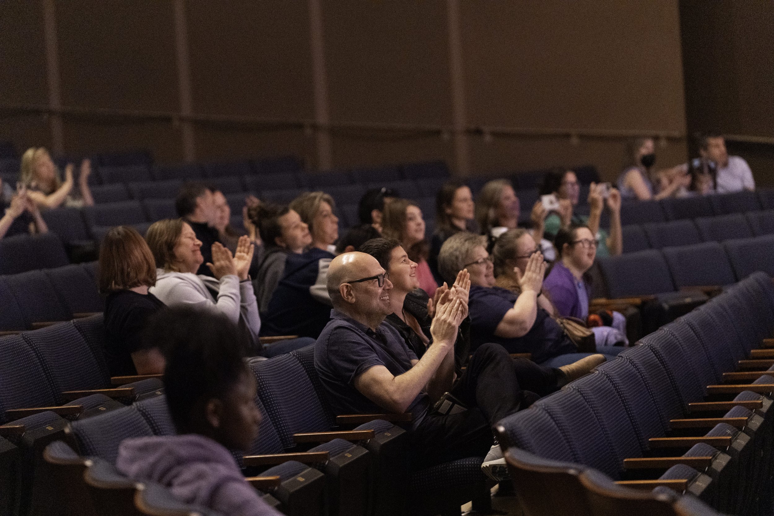 Audience members seated in a theater, clapping and smiling during a performance or presentation.