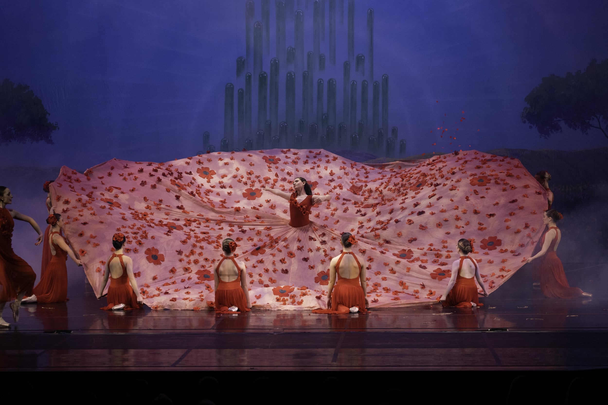Ballet dancers perform on stage, with a central dancer holding a large pink fabric decorated with red flowers, surrounded by others in red dresses.