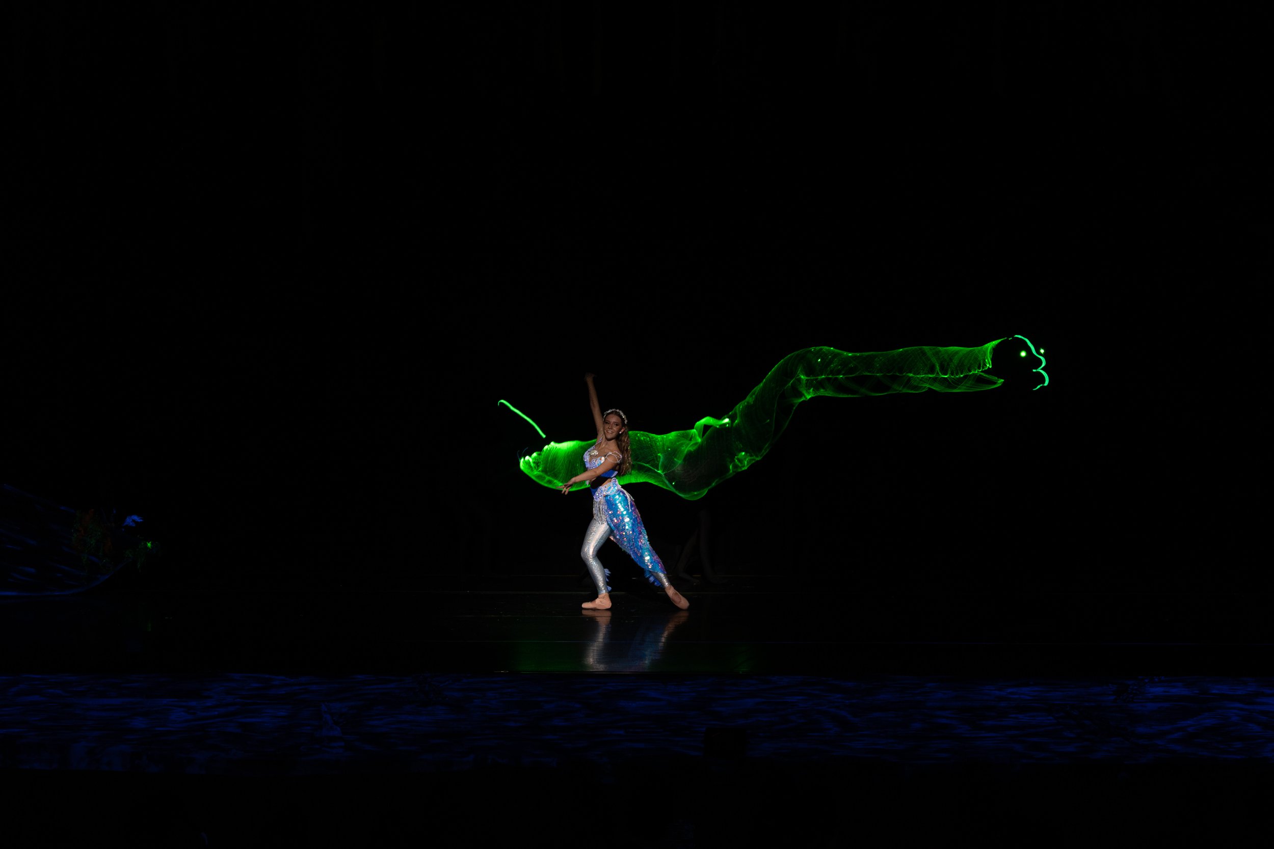 A performer in a shiny costume dances on stage with a green light trail resembling a dragon or serpent in the dark background.