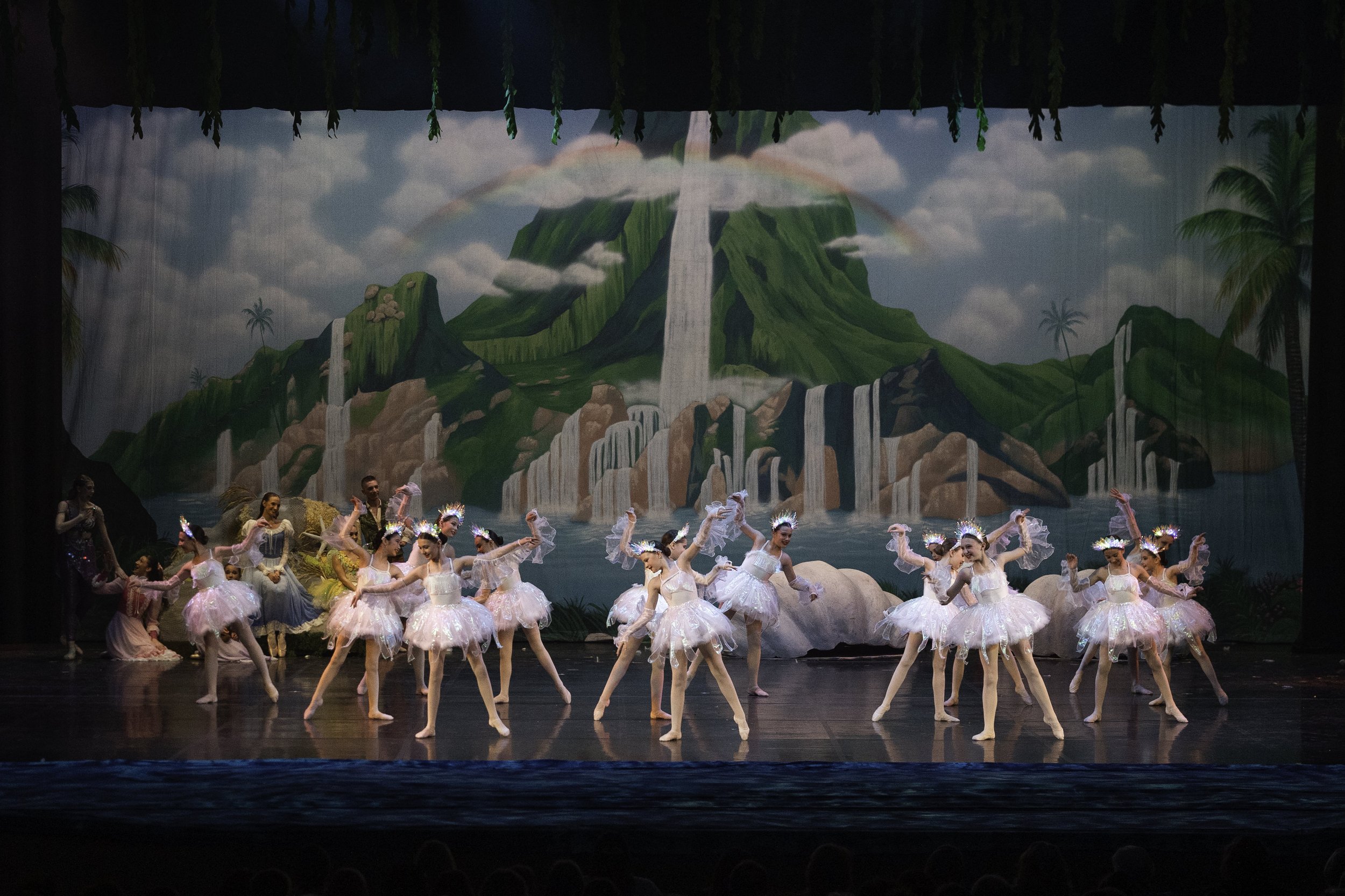 Ballet dancers dressed as fairies on stage with a background of a painted waterfall, mountain, and tropical trees.