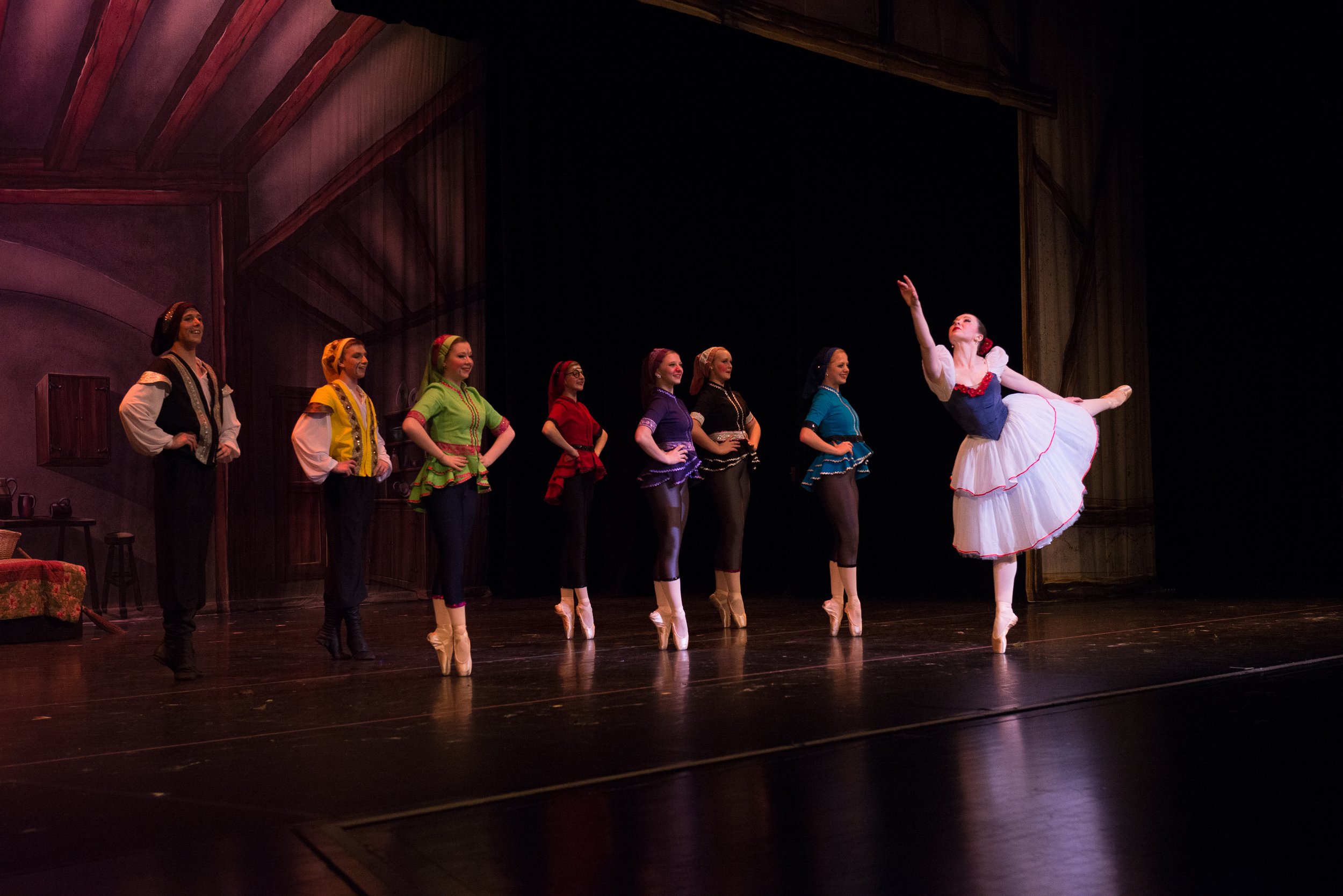 Ballet dancers performing on stage with one dancer in a white tutu and black vest executing a pointe move, while seven other dancers in colorful costumes observe.