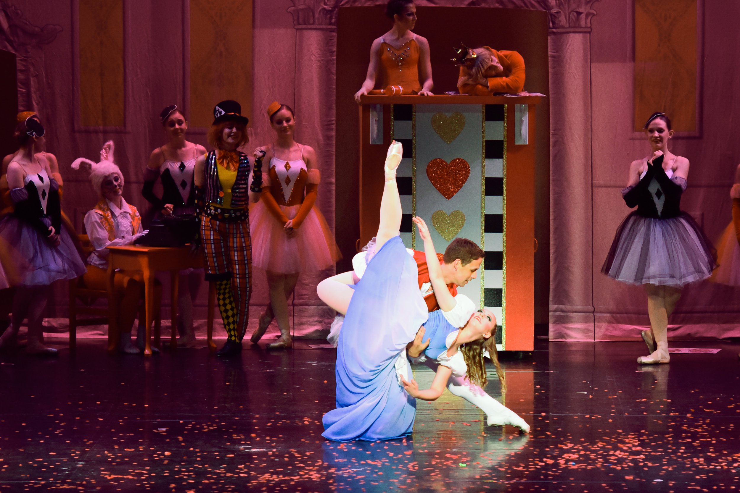 Two ballet dancers performing on stage, with one holding the other in a dip. The stage is decorated with a whimsical, colorful set featuring a tall structure with hearts and stripes, and several performers dressed in colorful costumes and tutus watch