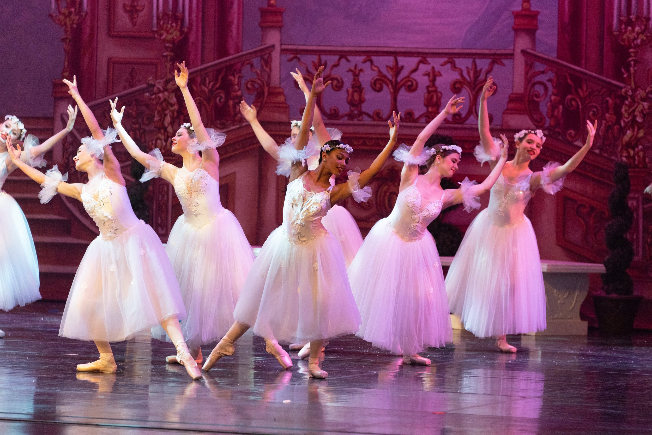 Ballet dancers in white tutus performing on stage with pink and purple backdrop.