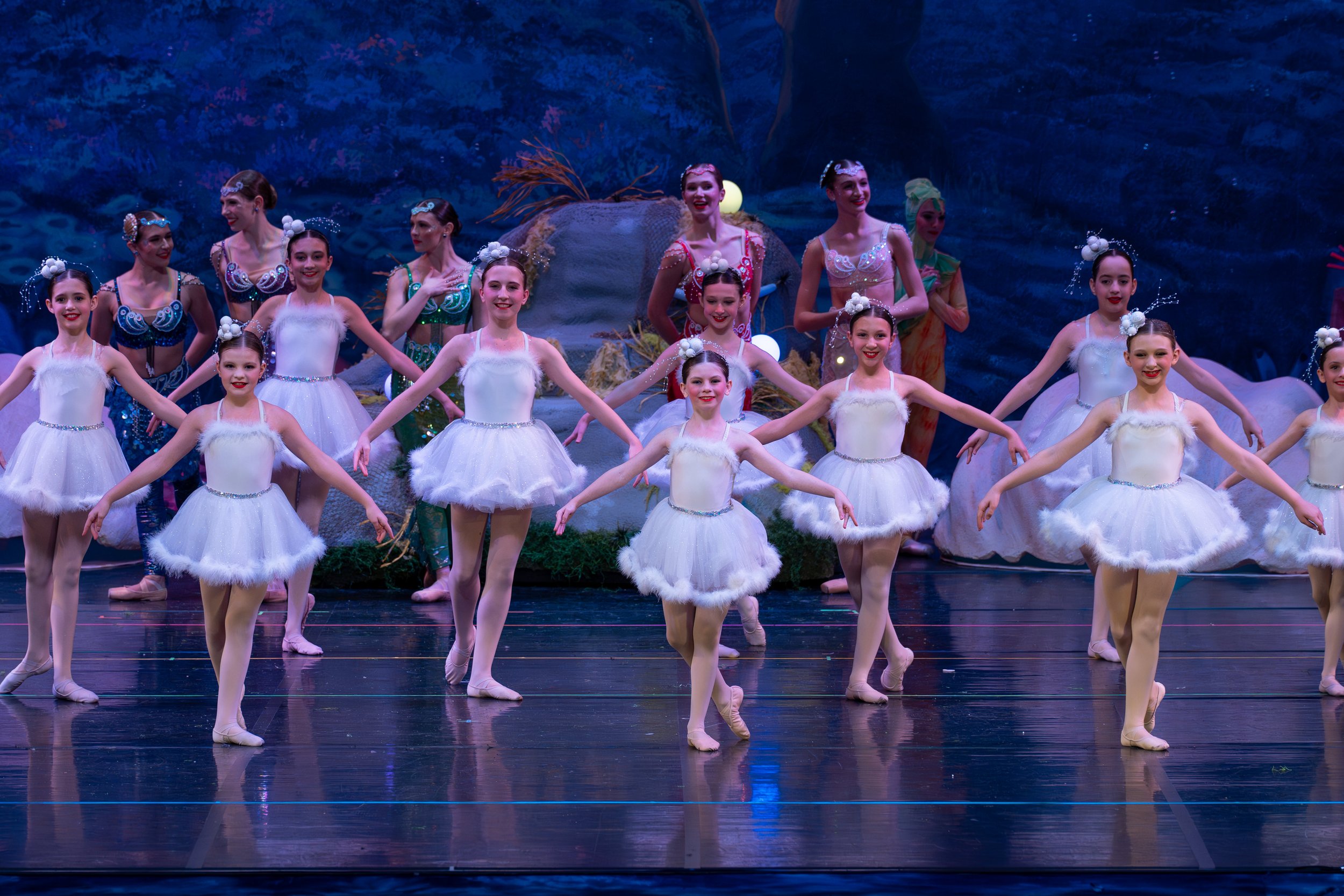 A group of young ballet dancers performing on stage, dressed in white tutus and leotards, with some adult dancers in colorful costumes in the background.