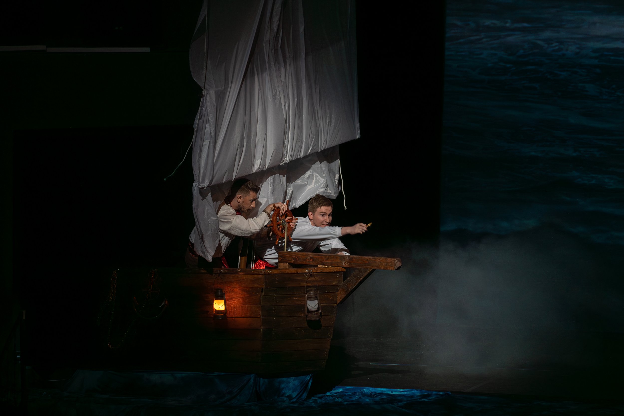 Actors dressed as pirates on a stage with a dark water backdrop, one man steering and the other pointing forward from a wooden pirate ship prop.