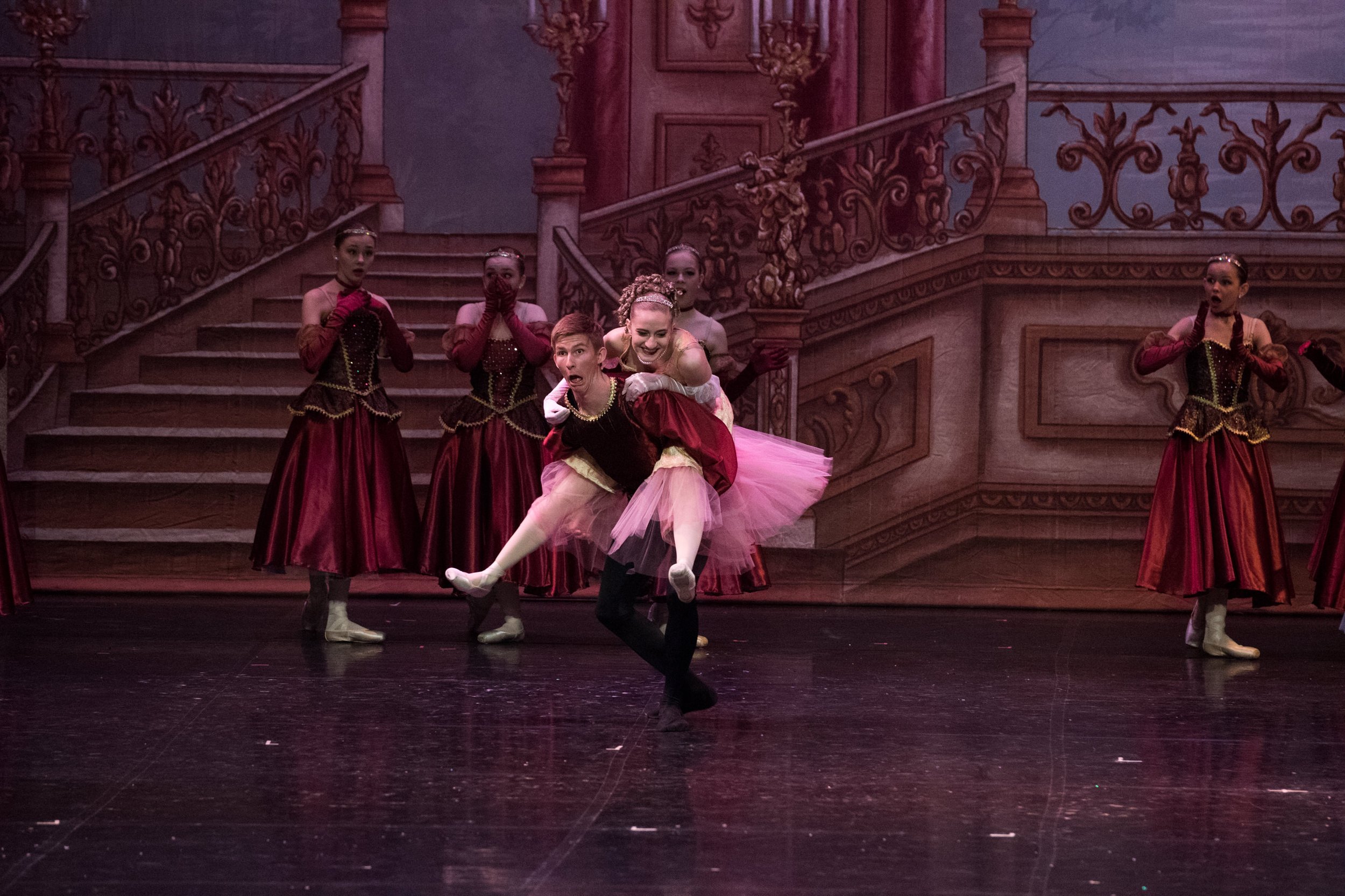 Ballet dancers perform on stage in front of a painted backdrop of a grand staircase and ornate walls, with one dancer lifting another in the foreground.