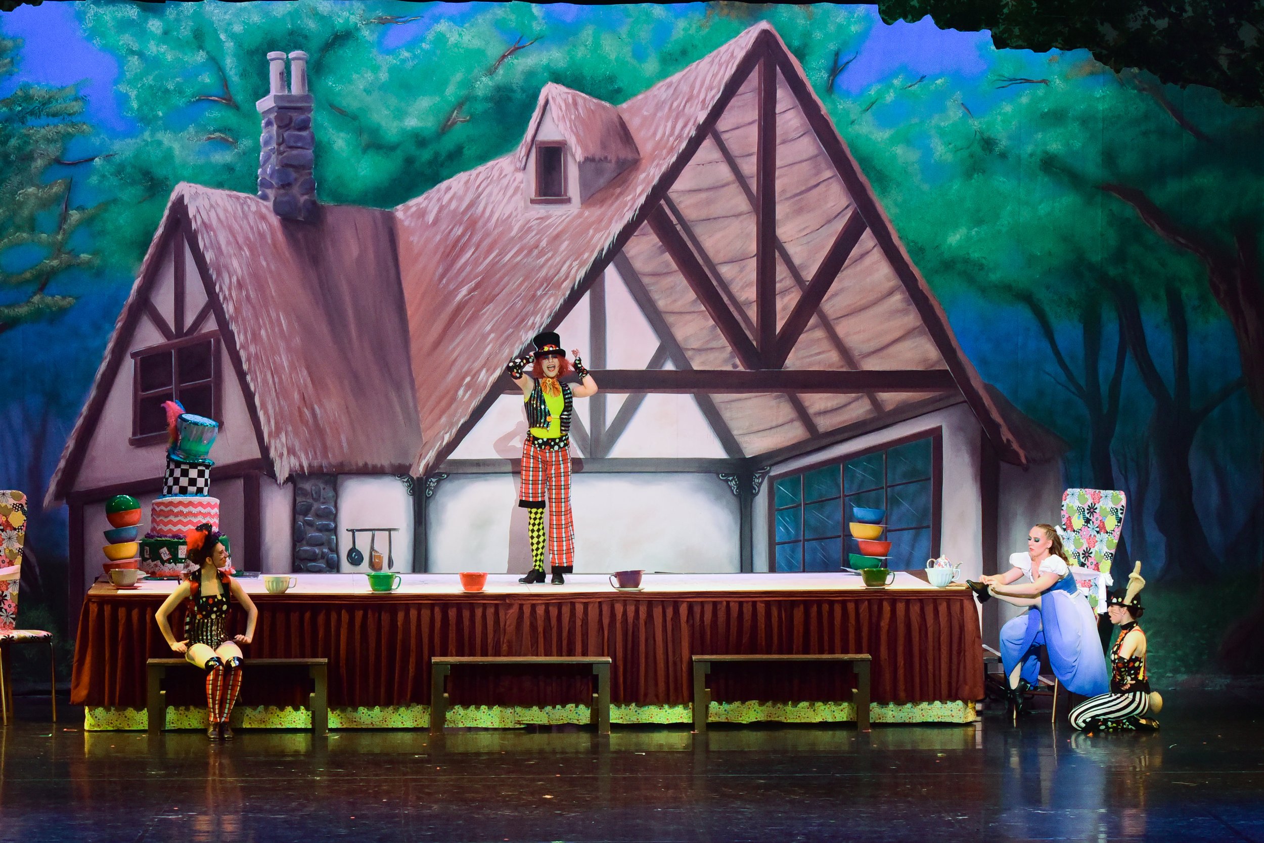 Stage scene of a theatrical performance with actors dressed as colorful clowns or jesters, a set resembling a house with a thatched roof, and a painted backdrop of trees and sky.