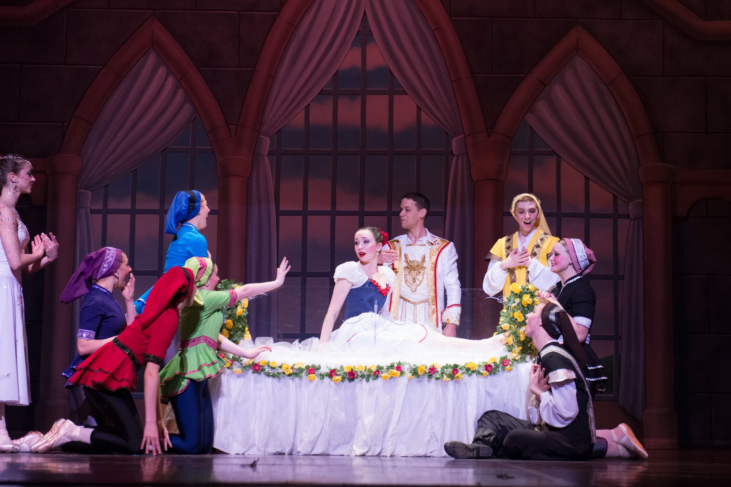 Scene from a theatrical performance showing a woman in a ball gown lying in a bed with several people around her, in a set resembling a castle chamber with large arched windows and velvet curtains.