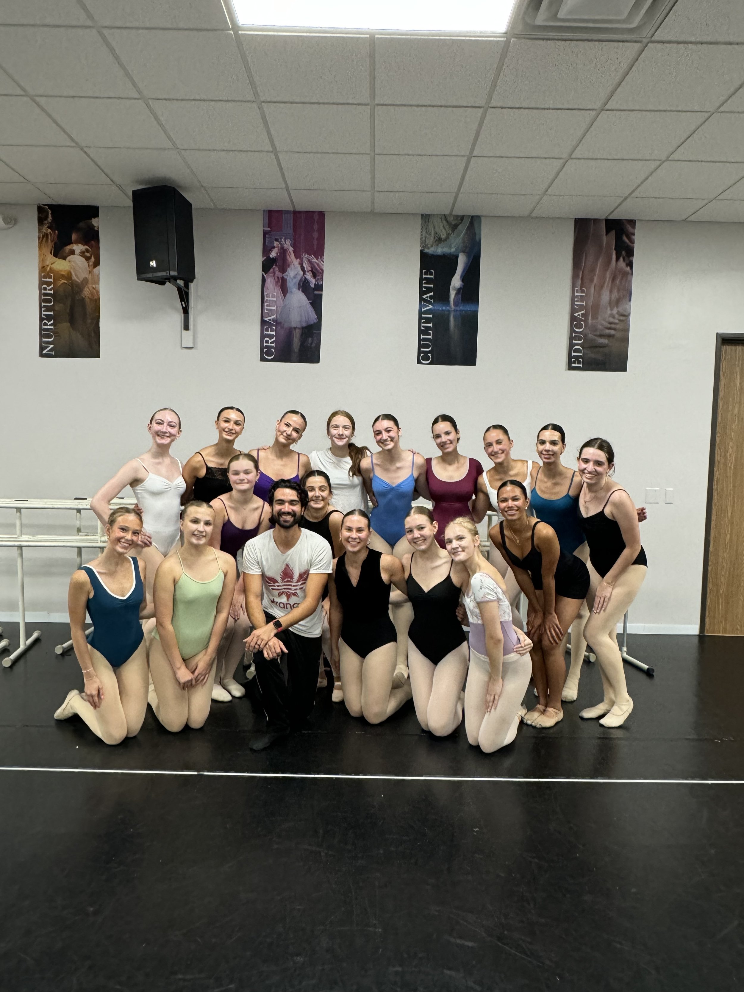Group of ballet dancers and instructor posing together in a dance studio, smiling. The dancers are wearing leotards and tights, and the studio has motivational banners with words: 'NUTURE', 'CREATE', 'CULTIVATE', 'EDUCATE' hanging on the wall.