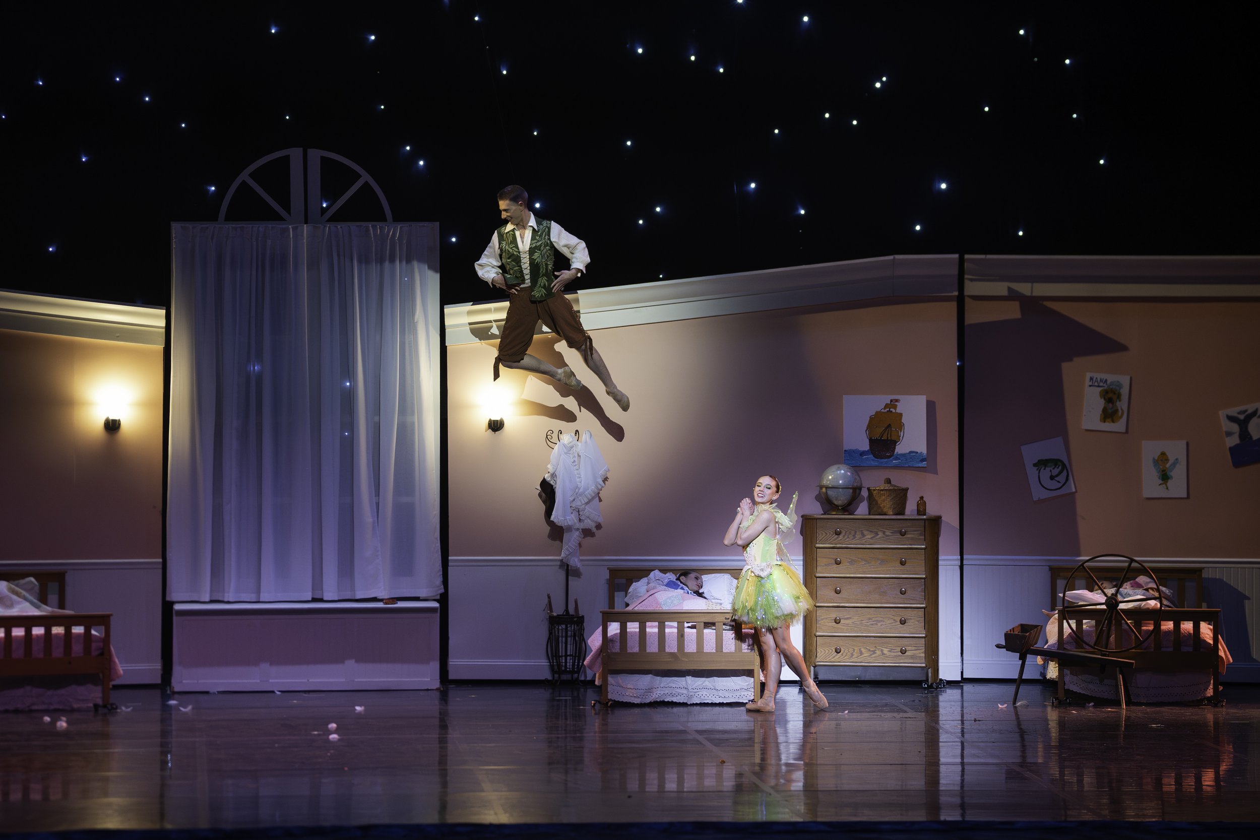 Stage scene from a ballet or theatrical production depicting three performers, including a male dancer mid-air, a female dancer on the floor, and a child in a bed, set in a colorful bedroom with beds, furniture, and wall art.