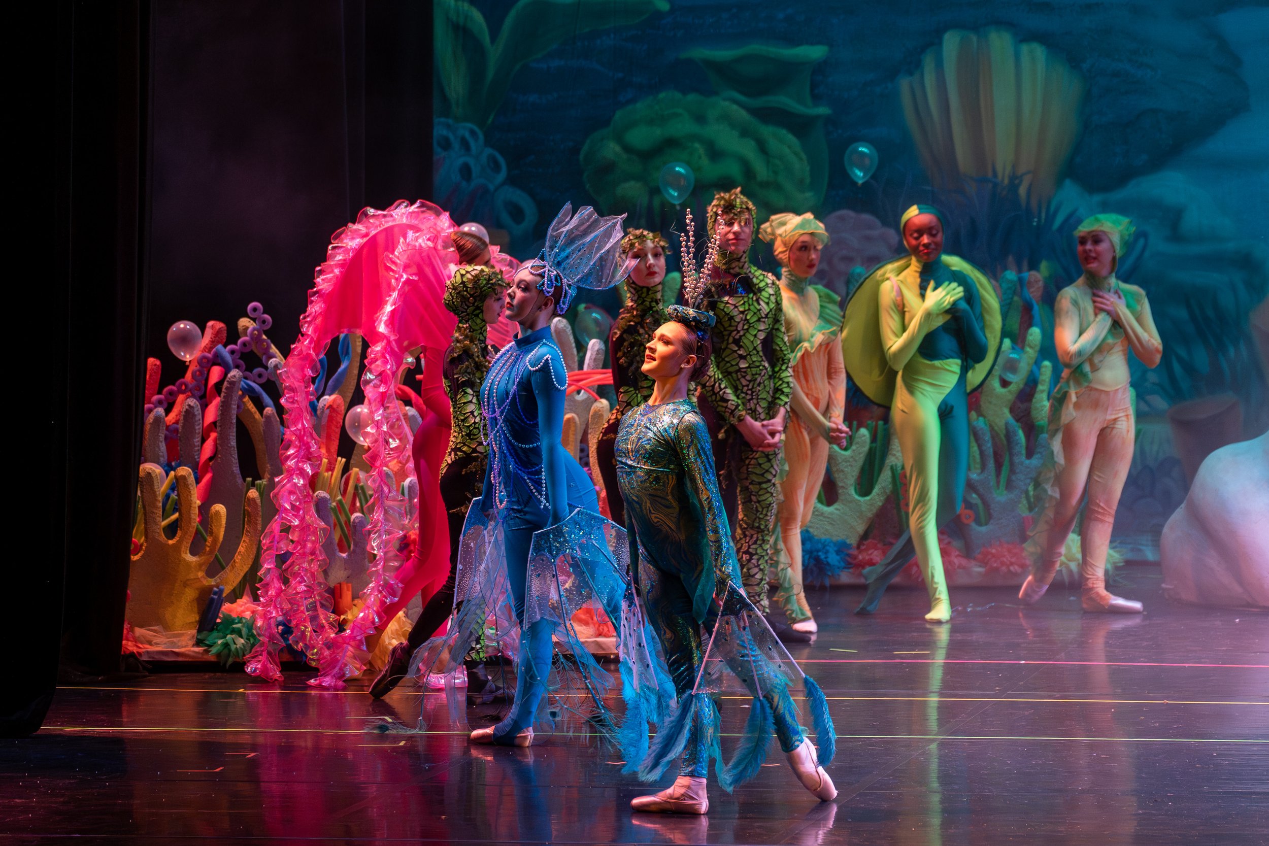 Ballet dancers in colorful costumes performing on stage with an undersea backdrop.