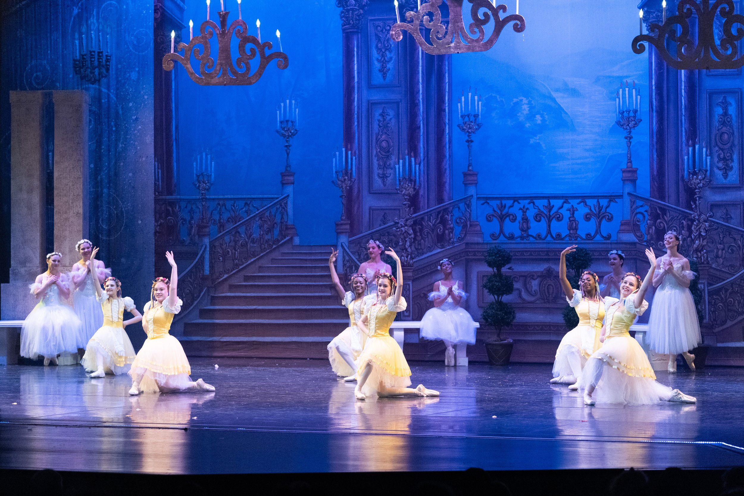 Ballet dancers performing on stage in white and yellow costumes in front of an ornate, castle-like backdrop with blue lighting and chandeliers.