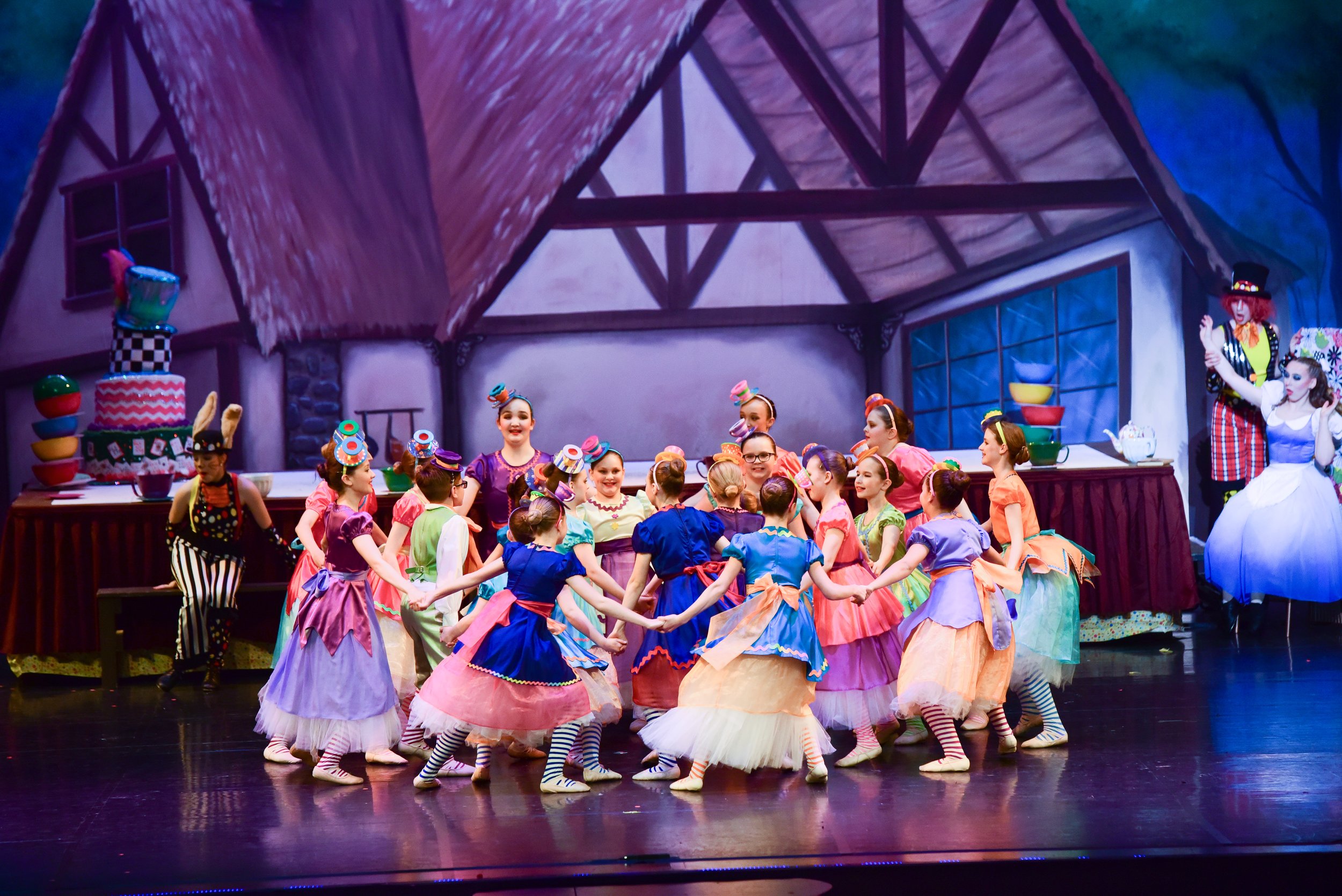 Children in colorful costumes dancing in a theatrical stage play with a backdrop of a whimsical kitchen scene.