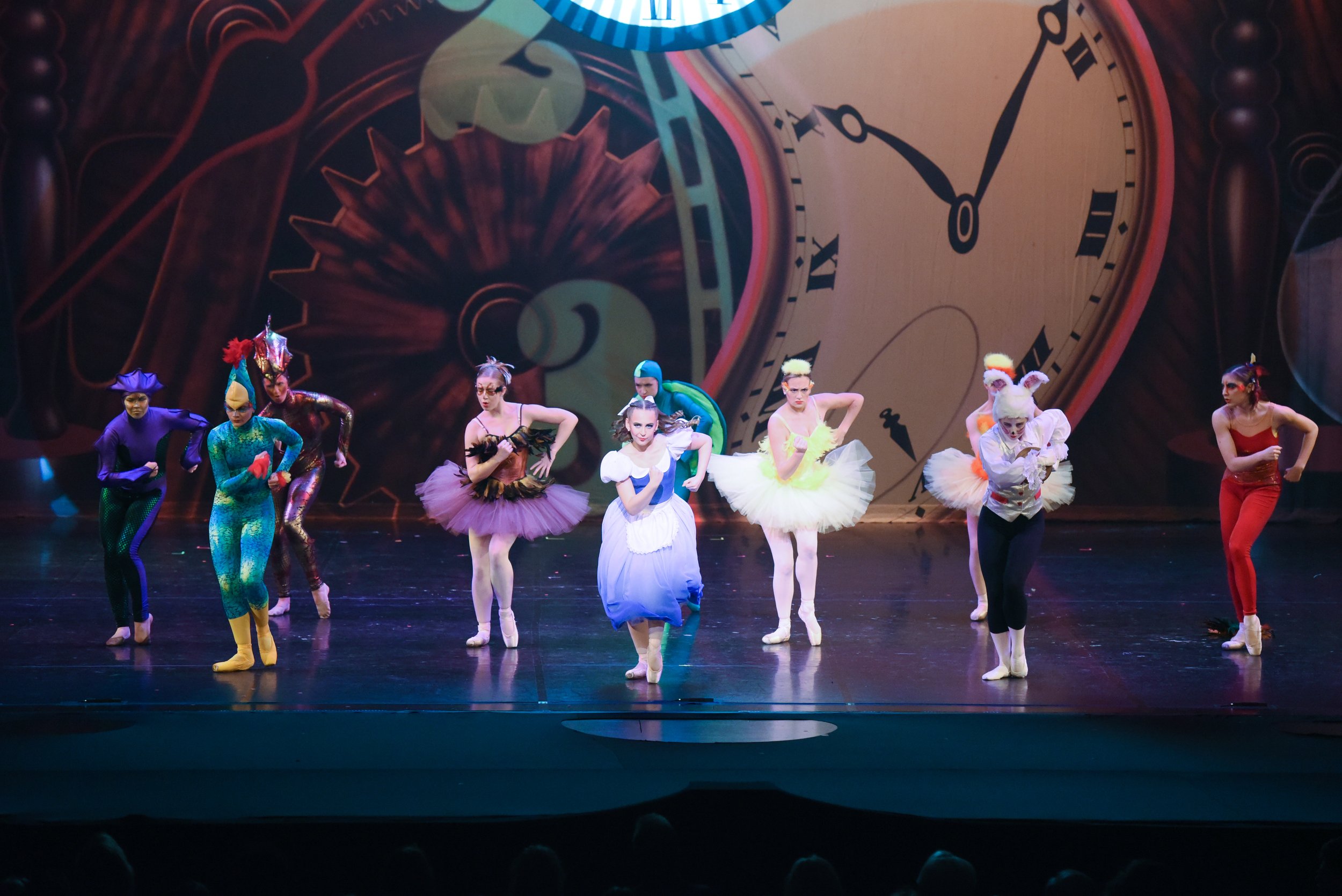 Ballet dancers dressed as characters from 'Alice in Wonderland' performing on stage with a large clock backdrop.