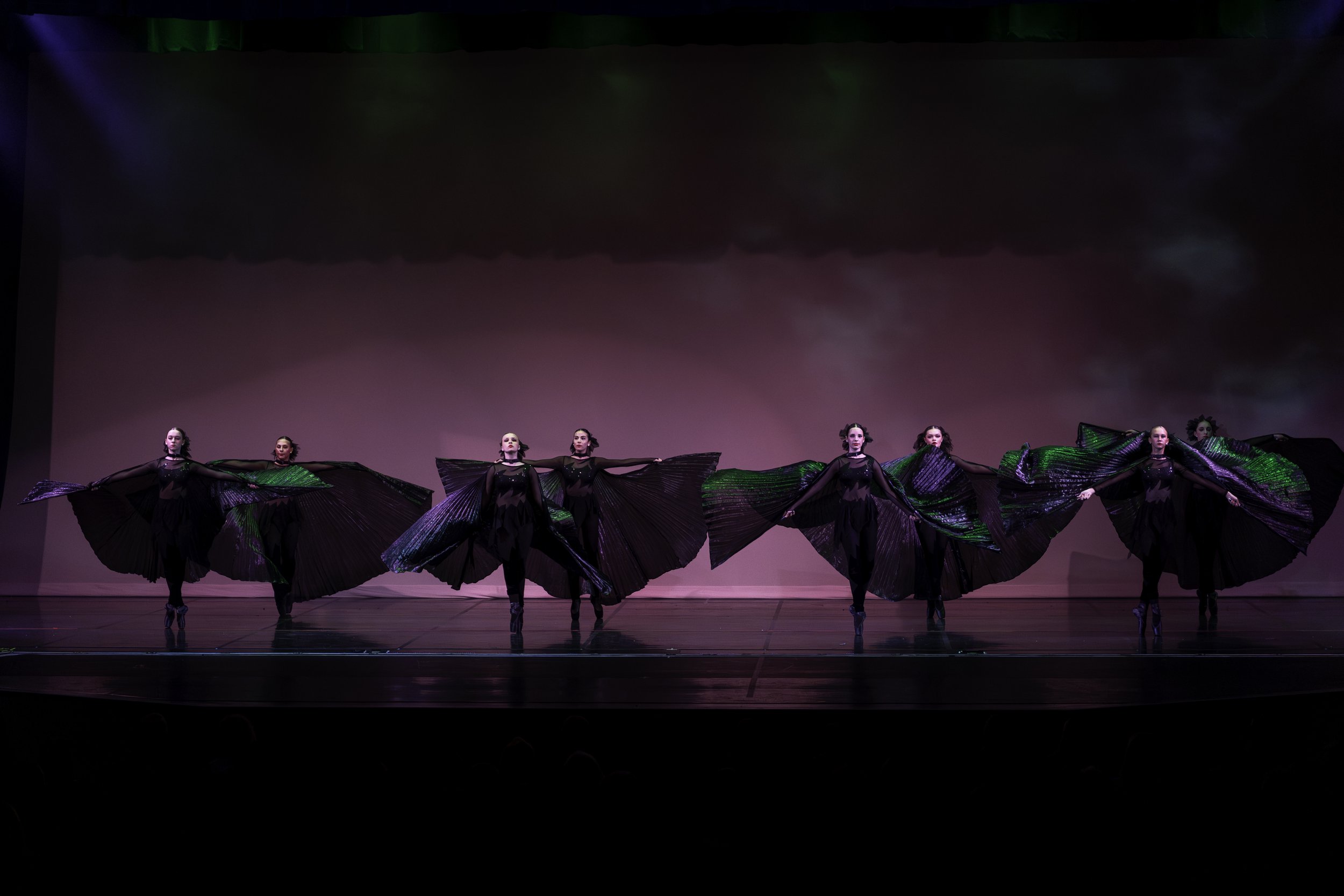 Six dancers in black costumes performing on stage with large pleated black and green wings, lit by colored stage lights, against a pink backdrop.