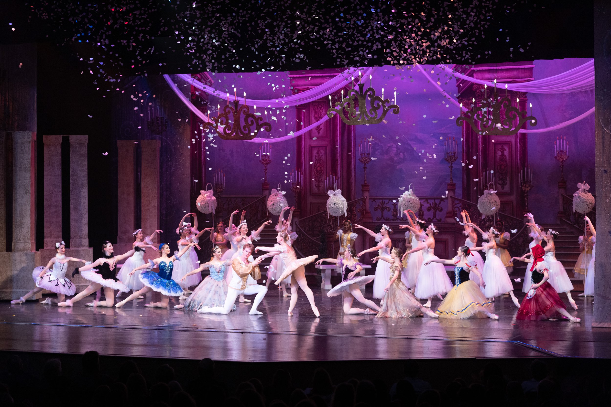 A ballet performance with multiple dancers wearing colorful costumes on a stage decorated to look like a palace, with chandeliers, draped fabric, and flower arrangements, under purple lighting.